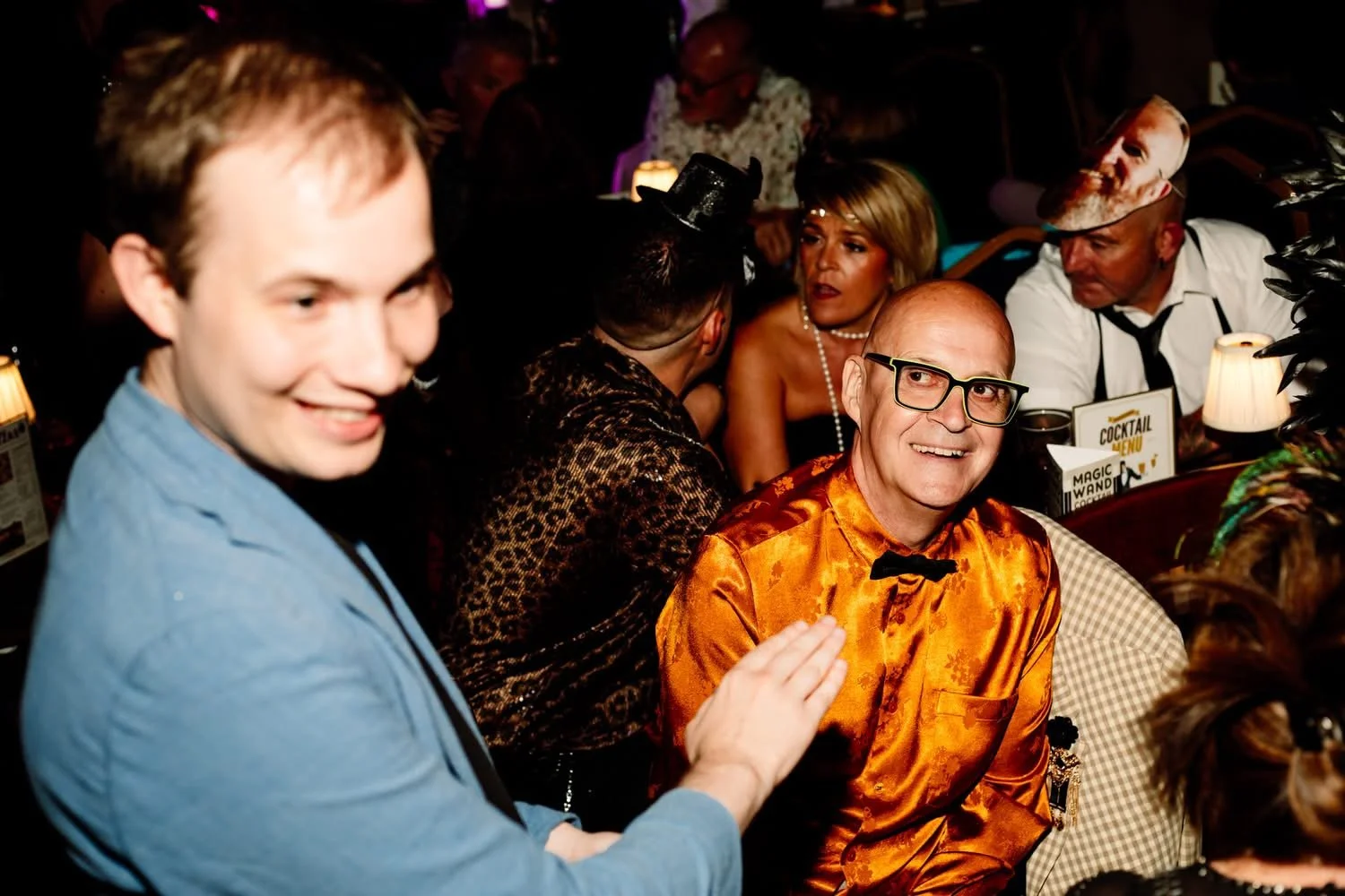 People socializing in a lively, dimly lit restaurant or bar, with some dressed in festive or vintage attire, including a man with glasses and a shiny orange shirt, and a woman in a string of pearls, sitting at a table with drinks and party menus.