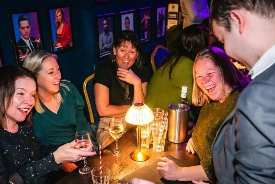 Four women laugh and enjoy drinks at a table as a restaurant magician entertains nearby. Photos are displayed on the wall behind them.