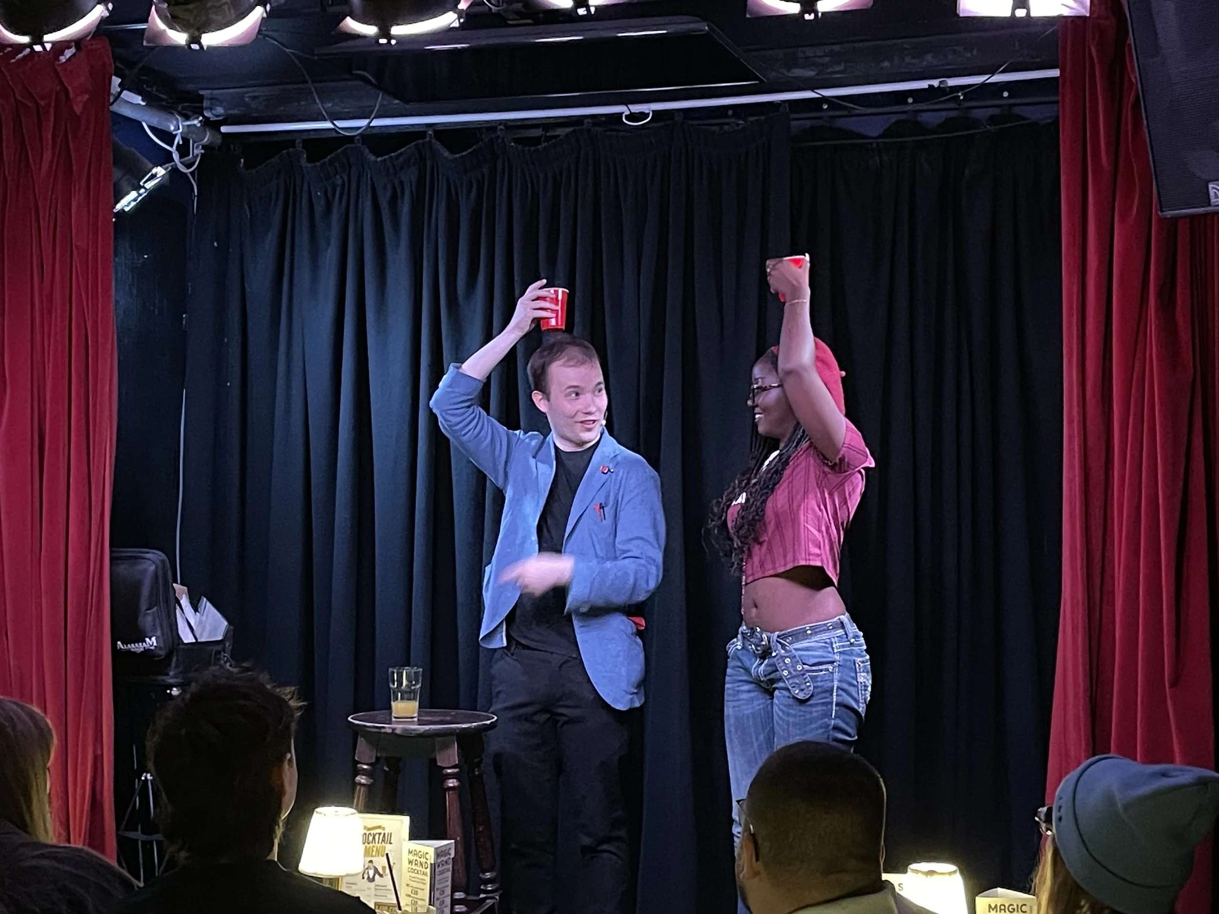 A man and woman on a stage with black curtains, each holding a red cup, engaging with an audience during a performance or comedy show, with tables and audience members visible in the foreground.