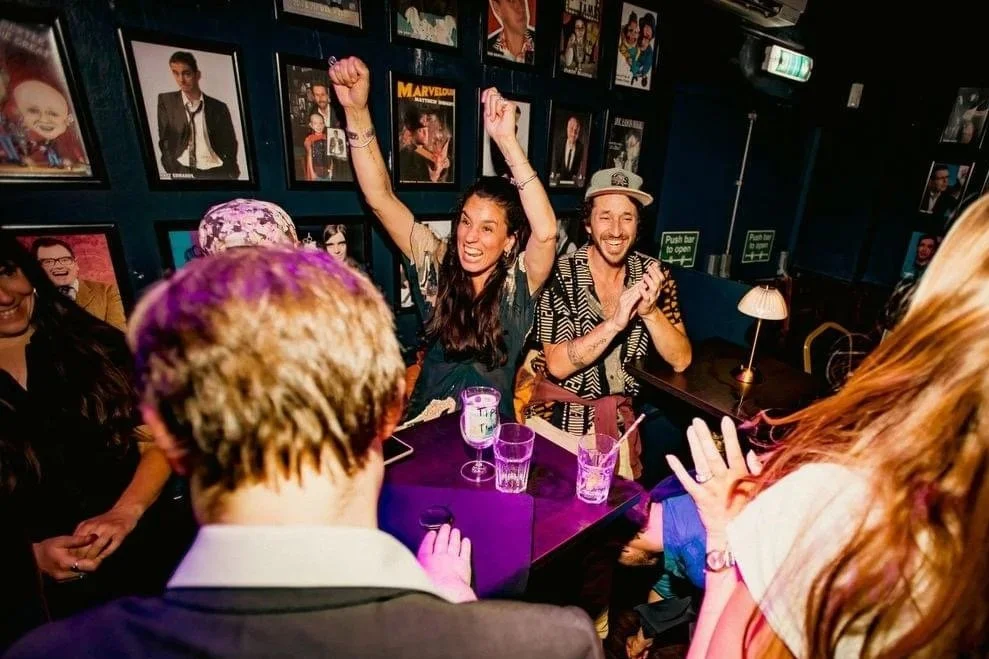 People cheer and clap as a magician in Weston-super-Mare performs at a lively, colorful bar with framed photos on dark blue walls.