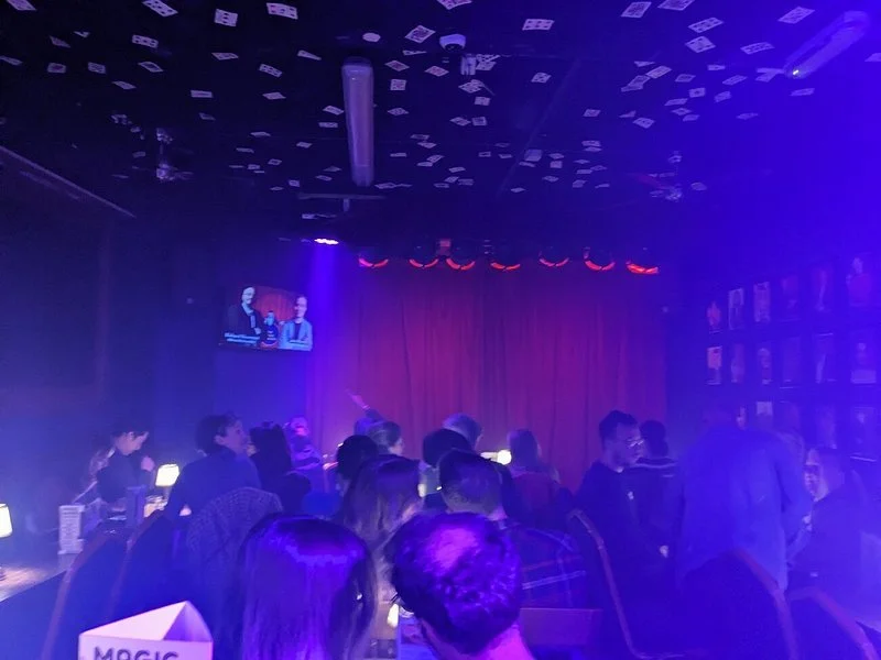 People sitting and standing at tables in a dark, blue-lit comedy club with a red curtain on stage and a large screen showing two people talking.