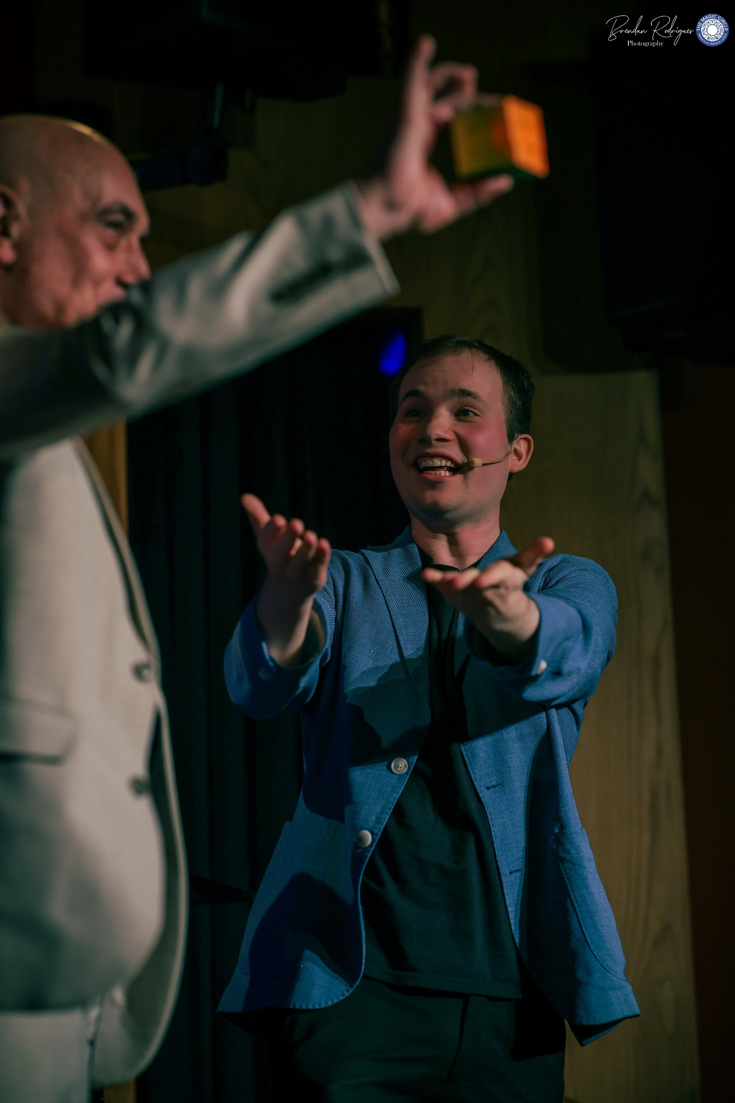 A young man in a blue blazer and black shirt, smiling and gesturing with his hands during a presentation or discussion, while an older man with a microphone headset, dressed in a white jacket, gestures with his hand, in a dimly lit room.