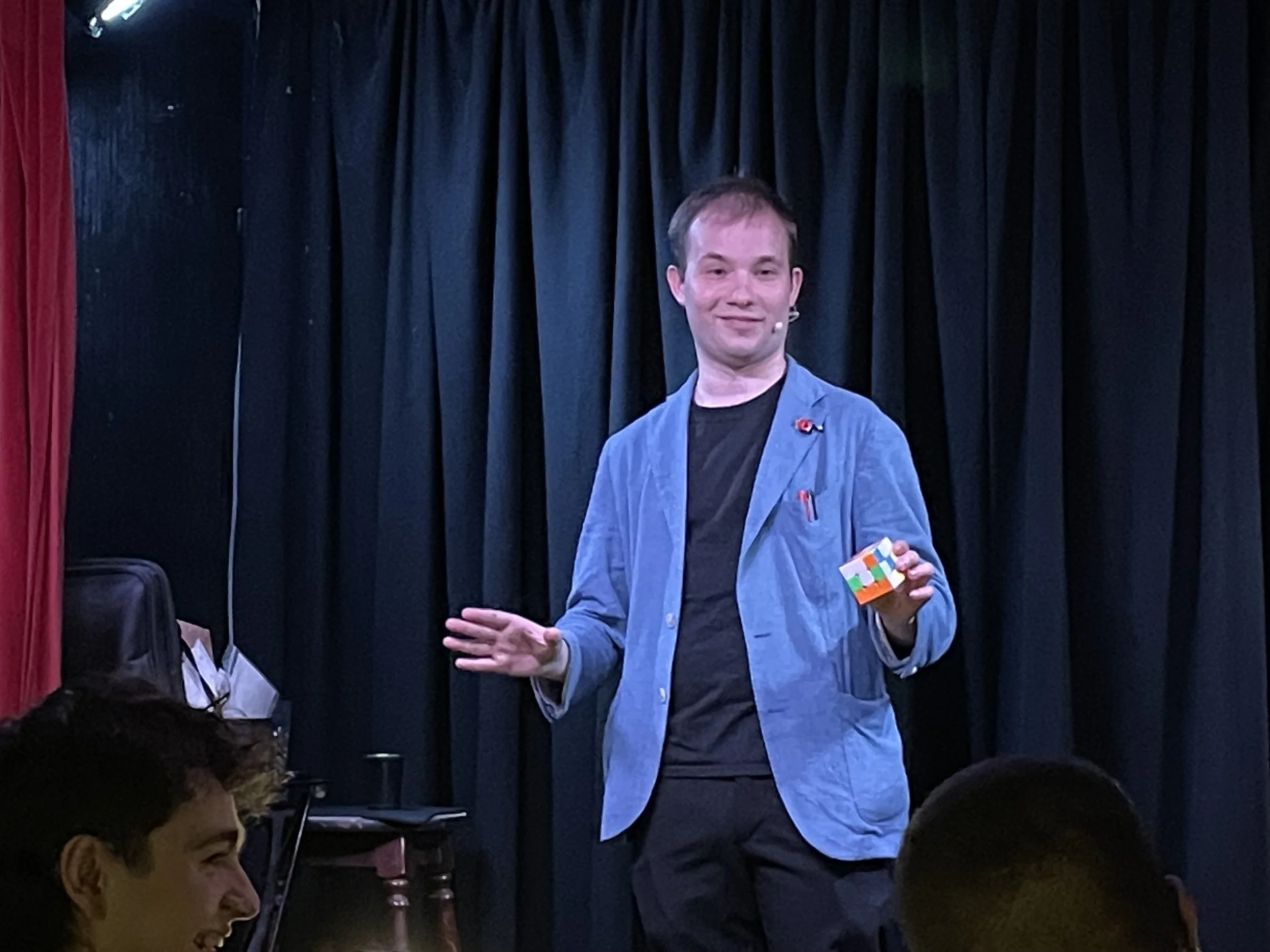 A man with light skin and short brown hair on stage, wearing a blue blazer over a black shirt, holding a colorful cube, with a microphone attached to his face, standing in front of black curtain.