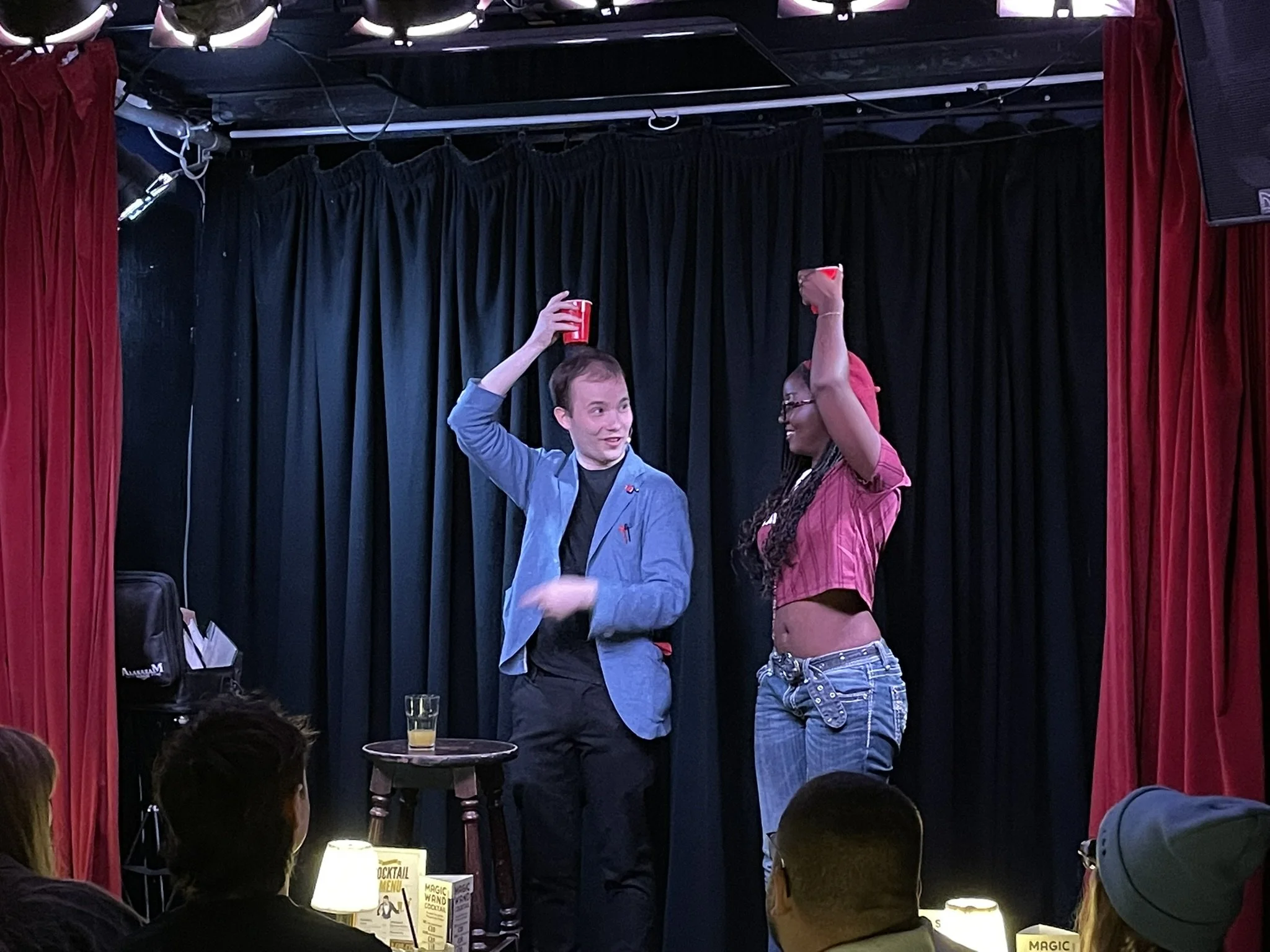 A man and a woman are on a small stage performing. They are holding red cups, with the man raising his cup while the woman is also raising hers. The audience is watching, and there are tables with drinks and napkins in front of them. The background i