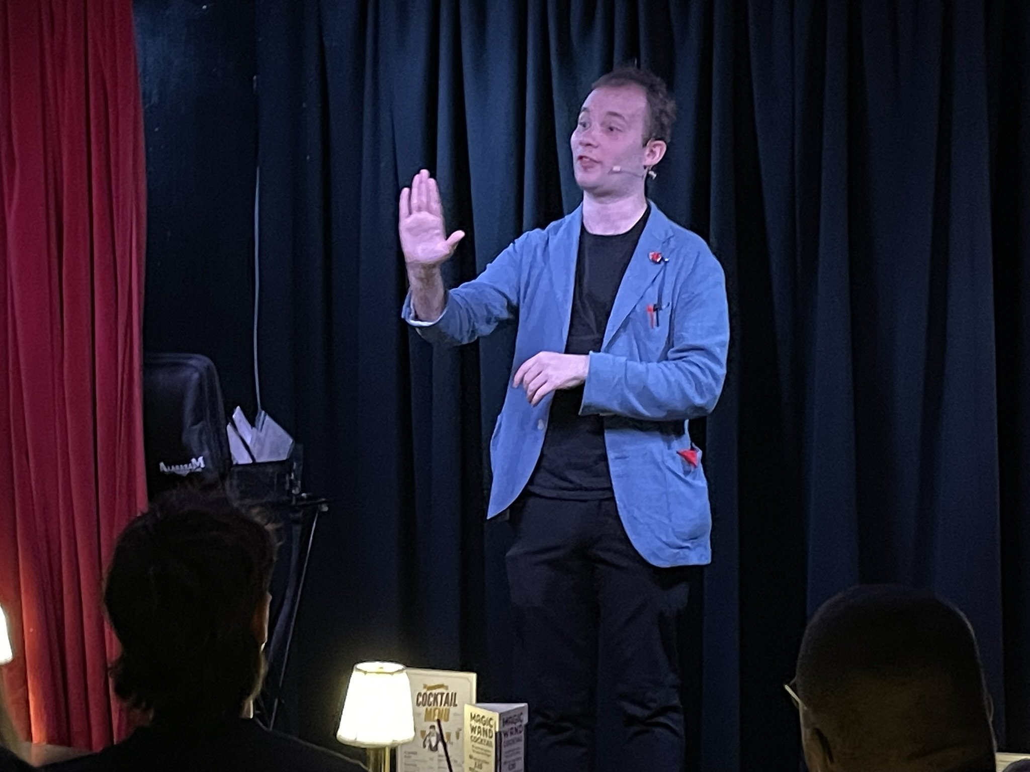 A man in a blue blazer giving a presentation on stage with black curtains behind him, and an audience with visible heads in the foreground.