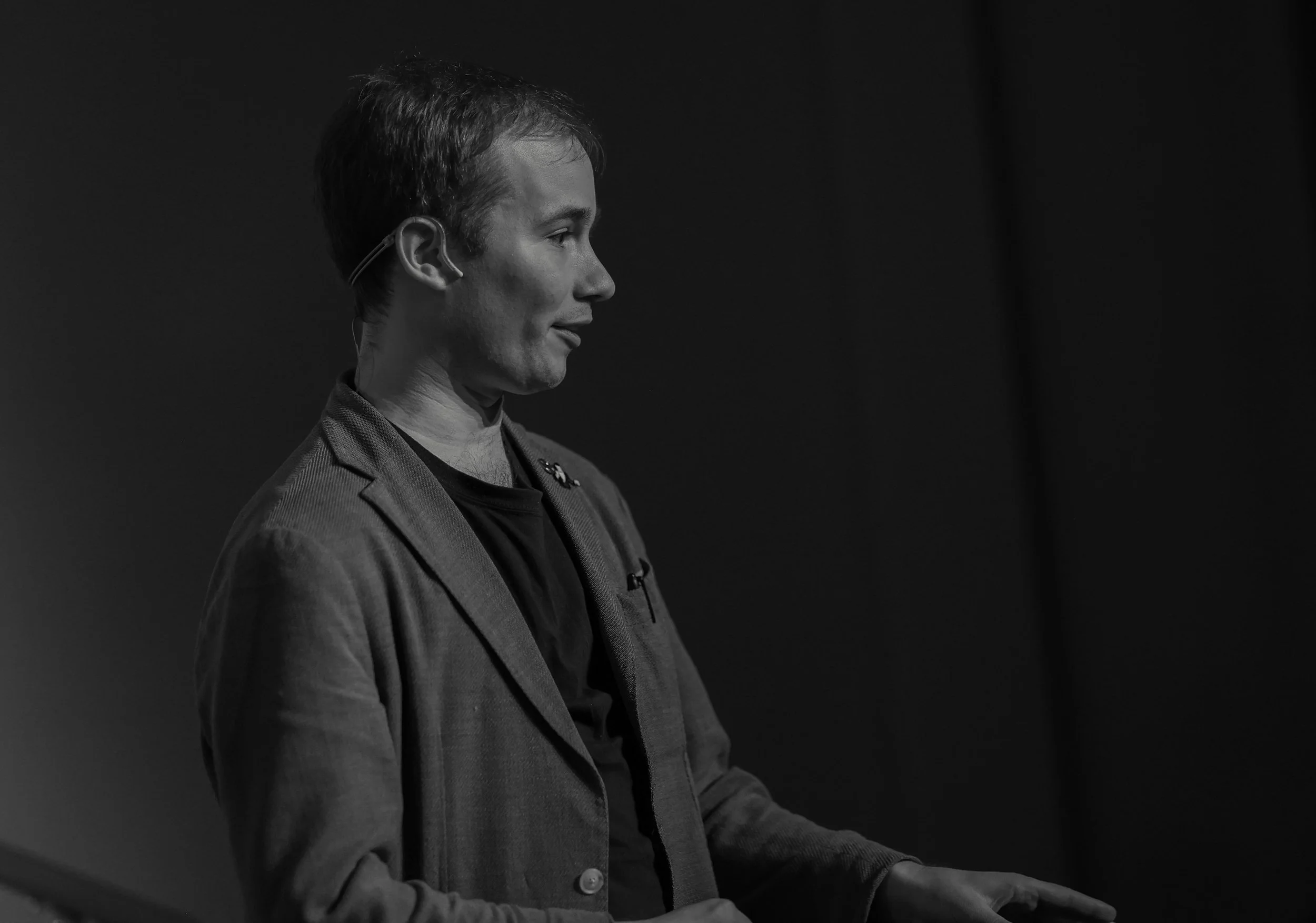 Side profile of a man with short hair wearing a blazer and a headset, standing against a dark background.