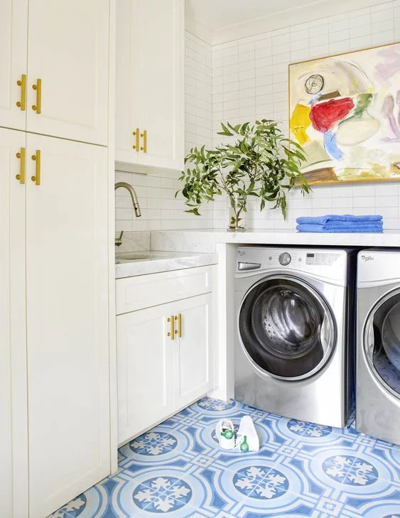 A blue tiled flooring in the laundry room balances with all-white cabinetry. Art pieces adorn the walls in multiple places throughout this home, but this is one of the most stunning. Abstract art is a great way to introduce color into a space.