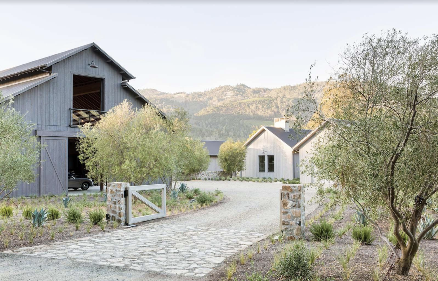 A welcoming entrance to the property is created using a simple stone and wood gate. As you look around, you’ll quickly notice that the spacious plot of land this home was built on includes not only an attached garage, but also a two-story barn for added storage. Both structures are cladded vertically, softening the facades and adding to their visual height. The designers and builders of this project collaborated with a dedicated landscaper to complete the stunning greenery surrounding the home which perfectly compliments the backdrop of rolling hills.