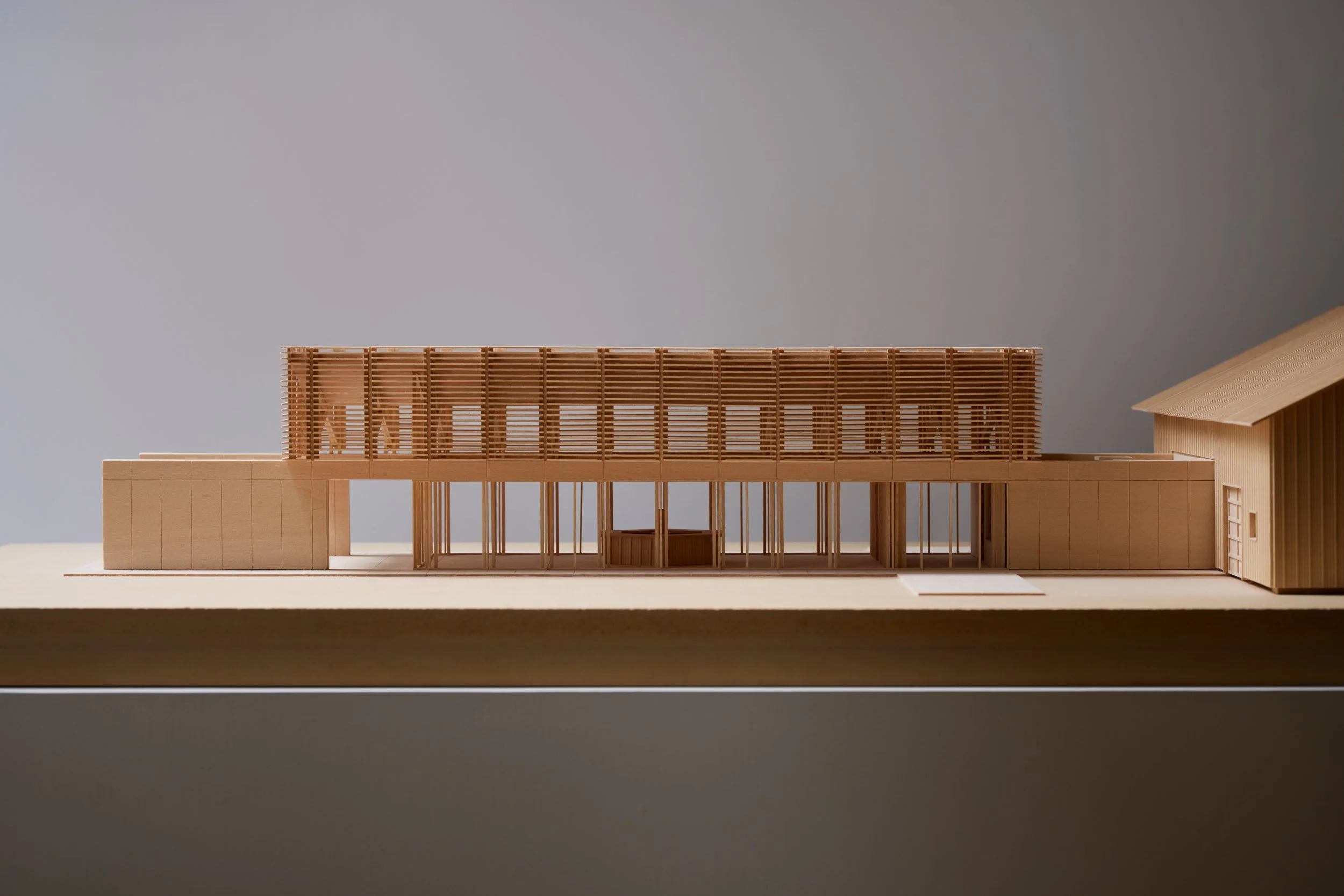 A detailed architectural model of a modern building, showcasing a structure with wooden slats and open spaces, placed on a stand against a plain background.
