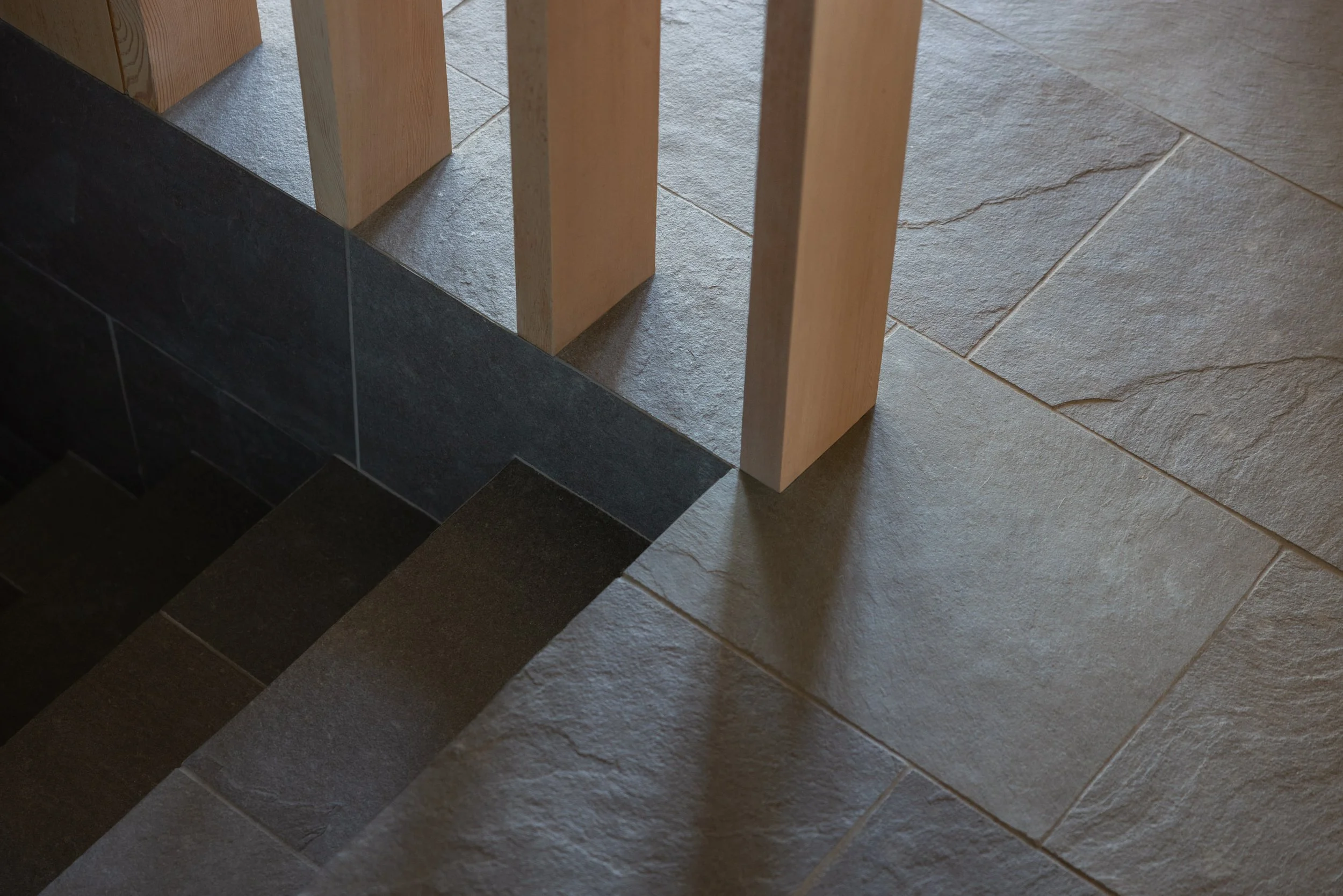 Close-up of a wooden chair leg on a tiled floor with different colored stone tiles and a shadow.