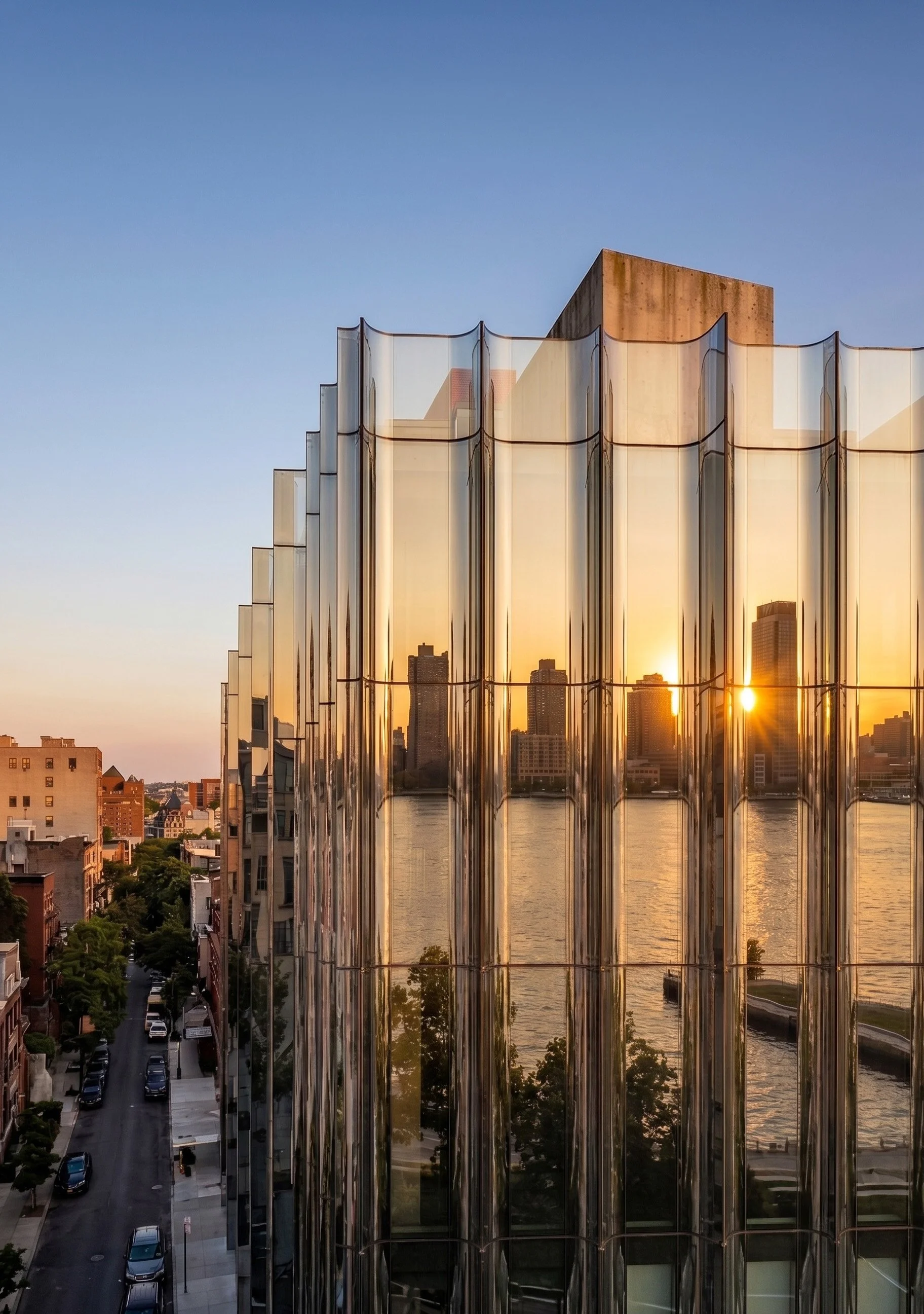 Sunset reflecting on a modern glass building with curved, vertical panes, overlooking a cityscape with high-rise buildings and a river.