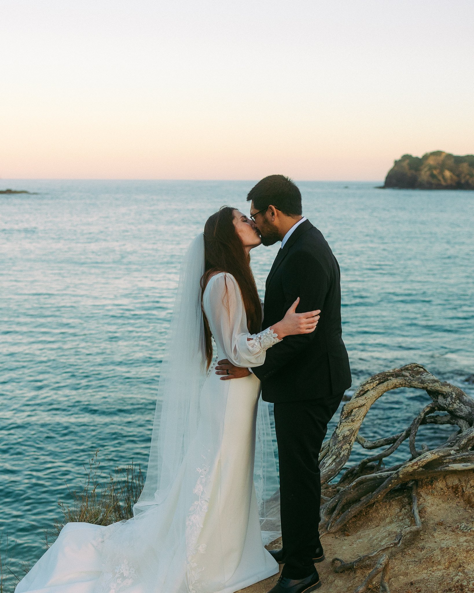 Wedding couple kissing by the ocean with sunset in the background.