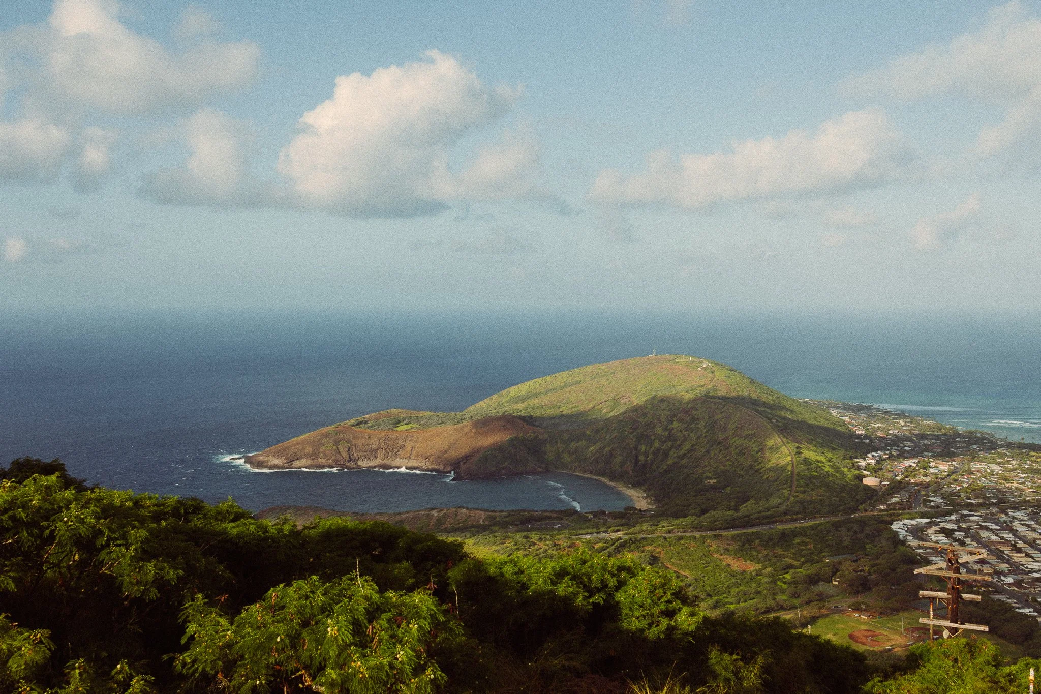 Scenic view of a coastal landscape in Oahu, Hawaii with a bay, green hills, and a town, under a partly cloudy sky.
