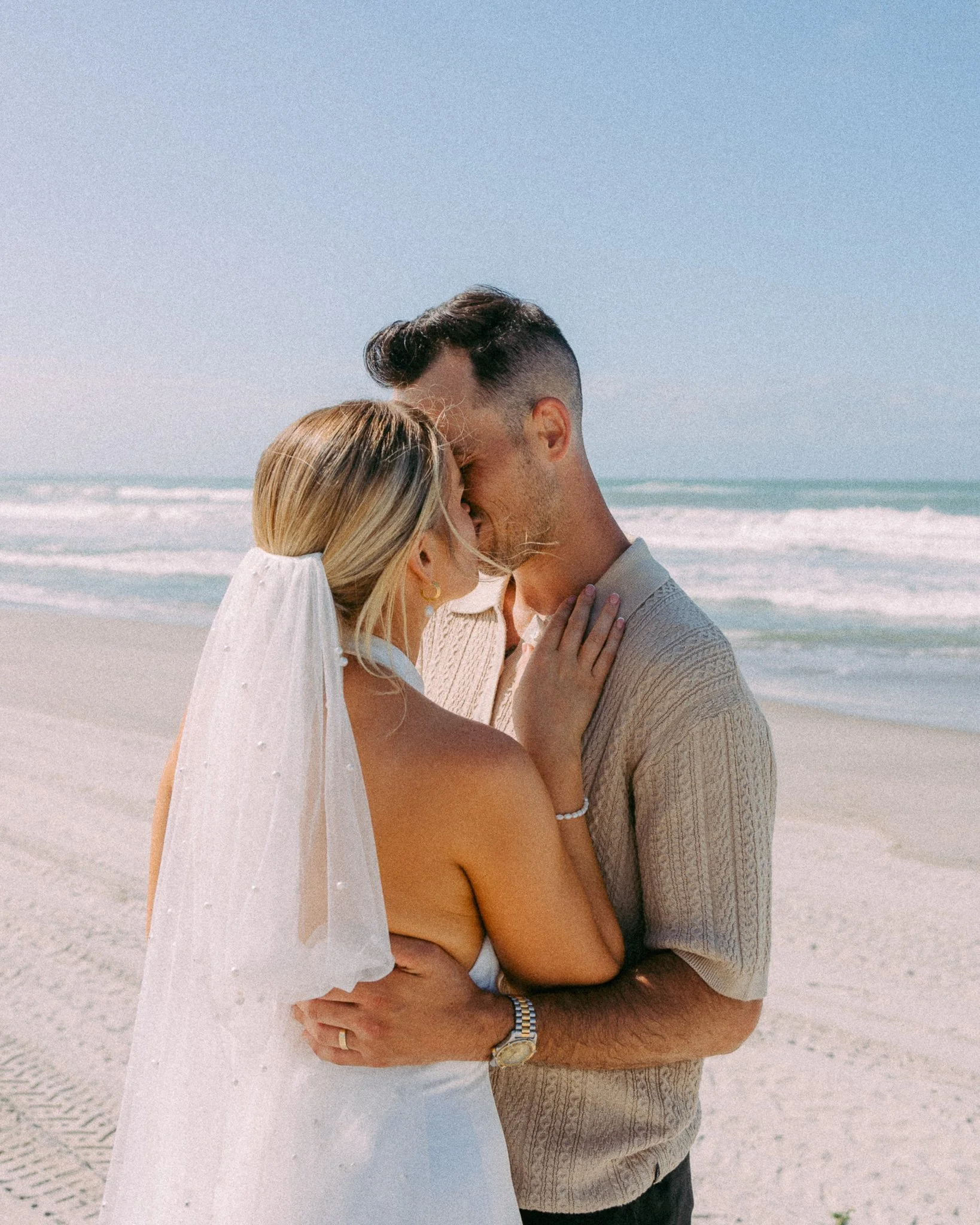 A bride and groom embrace on a beach, with the bride wearing a veil and the ocean in the background.