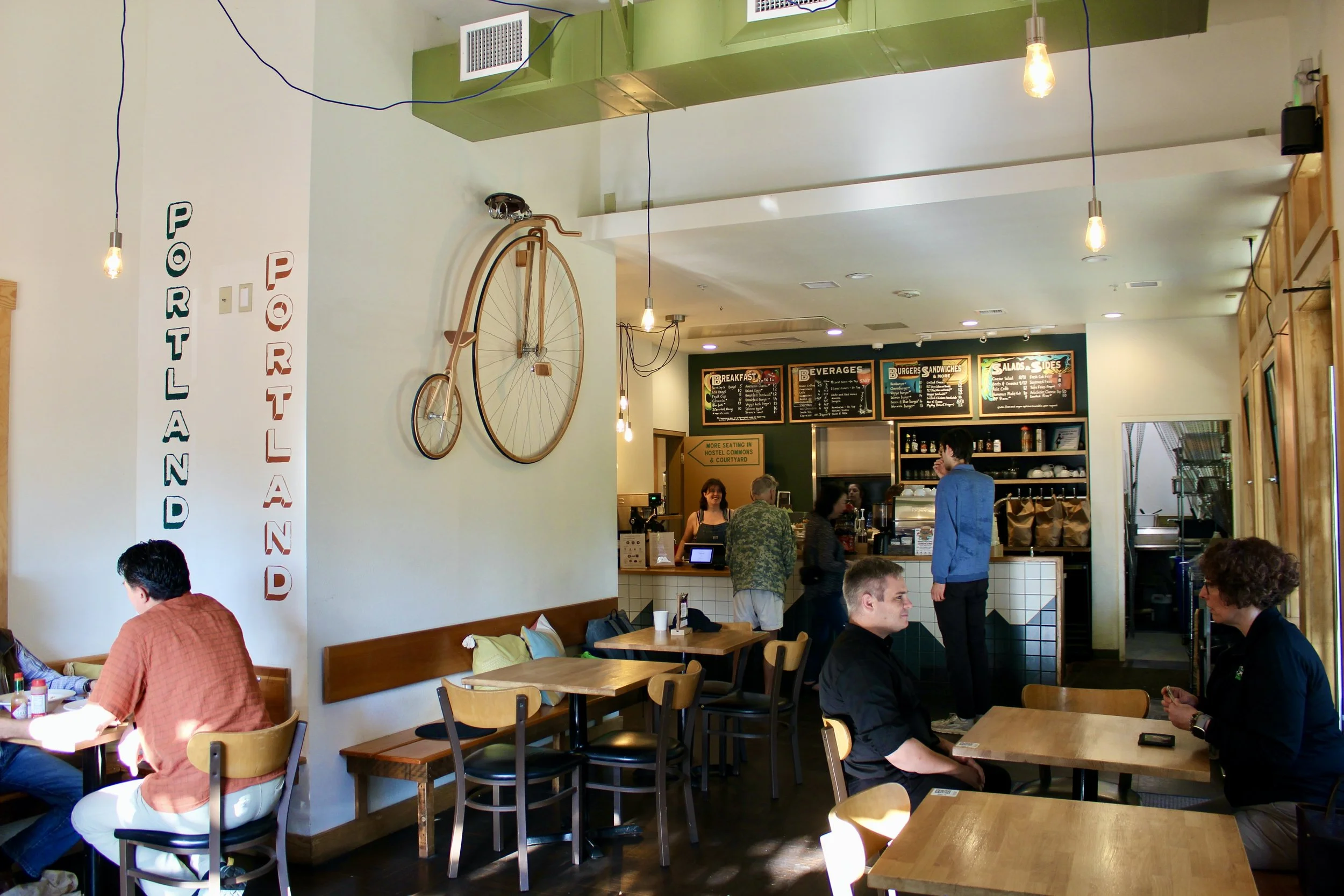Interior of Hostel Café with customers at wooden tables, chalkboard menu, and decorative bicycle on the wall.