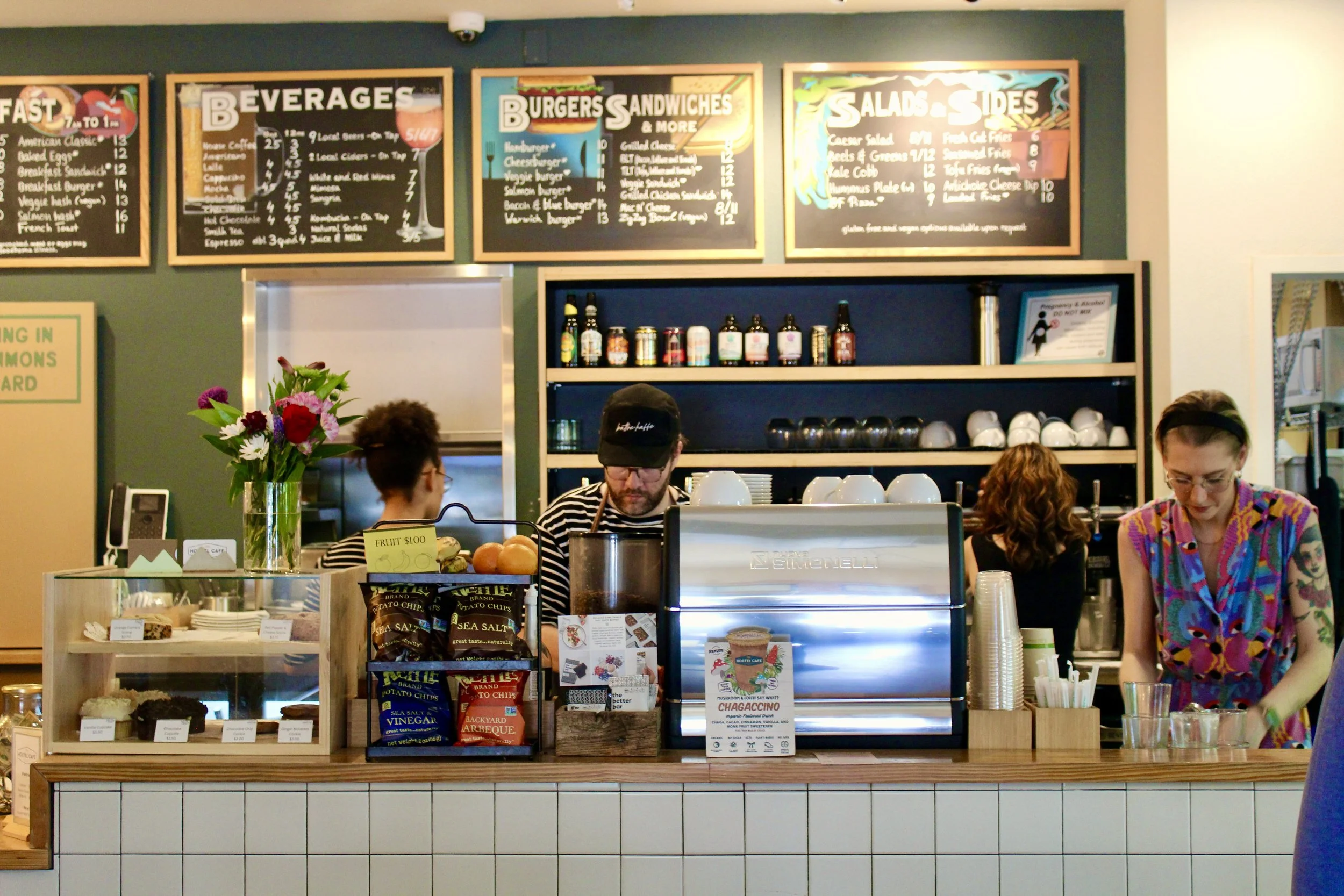 Inside the Hostel Café with barista preparing drinks, customers ordering, menu boards, flowers, and snacks on the counter machine.