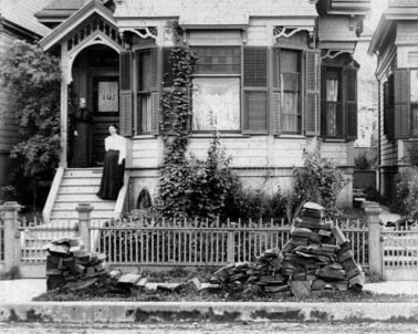 Black and white photo of a woman standing on the steps of a Victorian-style house with a small garden and a decorative fence in front.