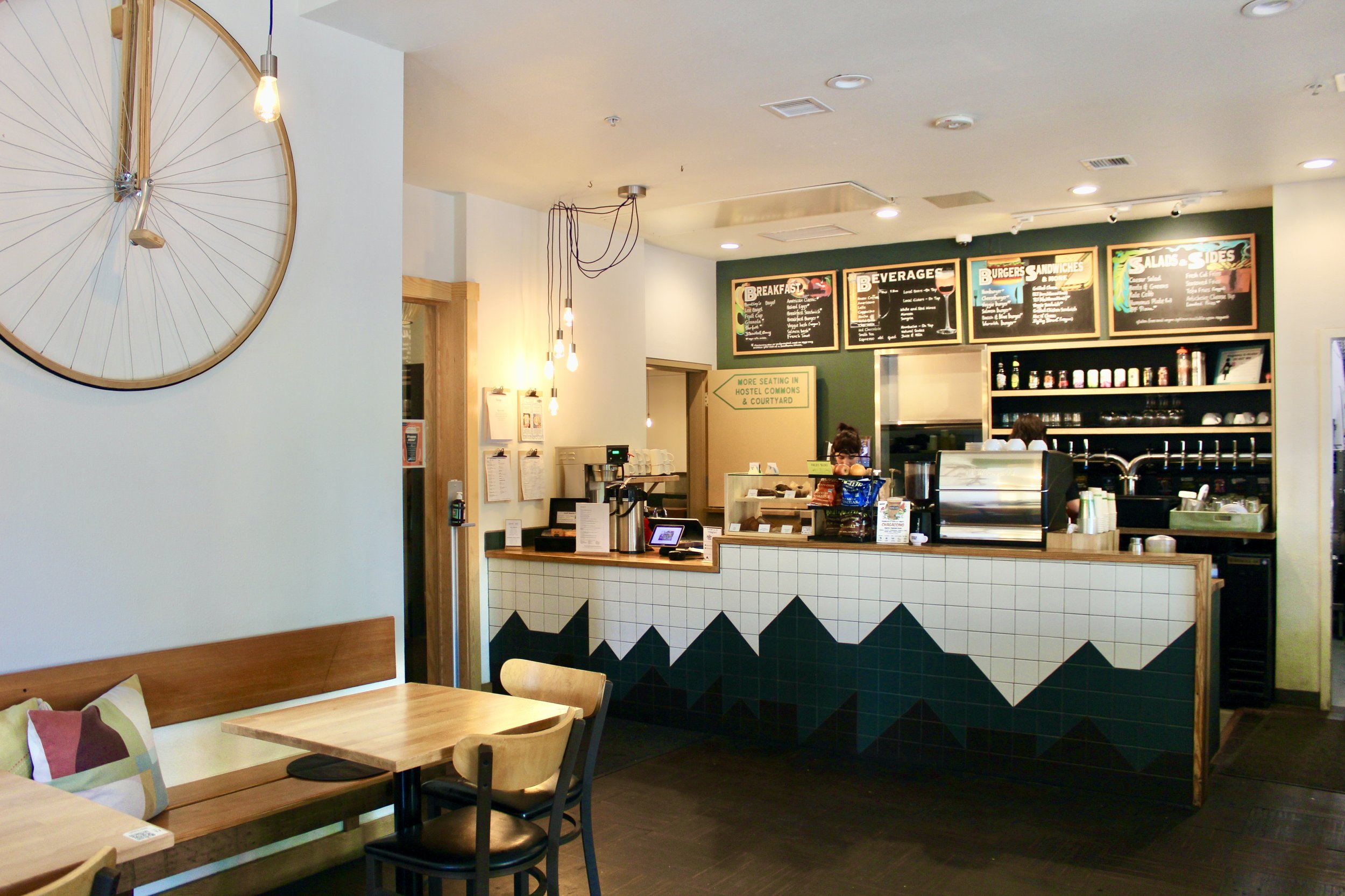 The Hostel Cafe interior with a front counter, menu boards, and a seating area with a bench and chairs. A large decorative bicycle wheel hangs on the white wall.