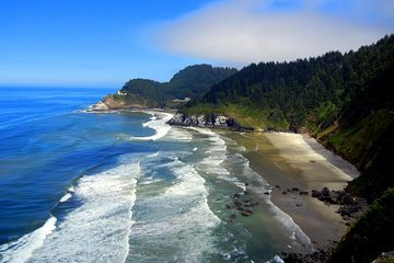 Coastal view of a sandy beach with waves, cliffs, and dense forested hills.
