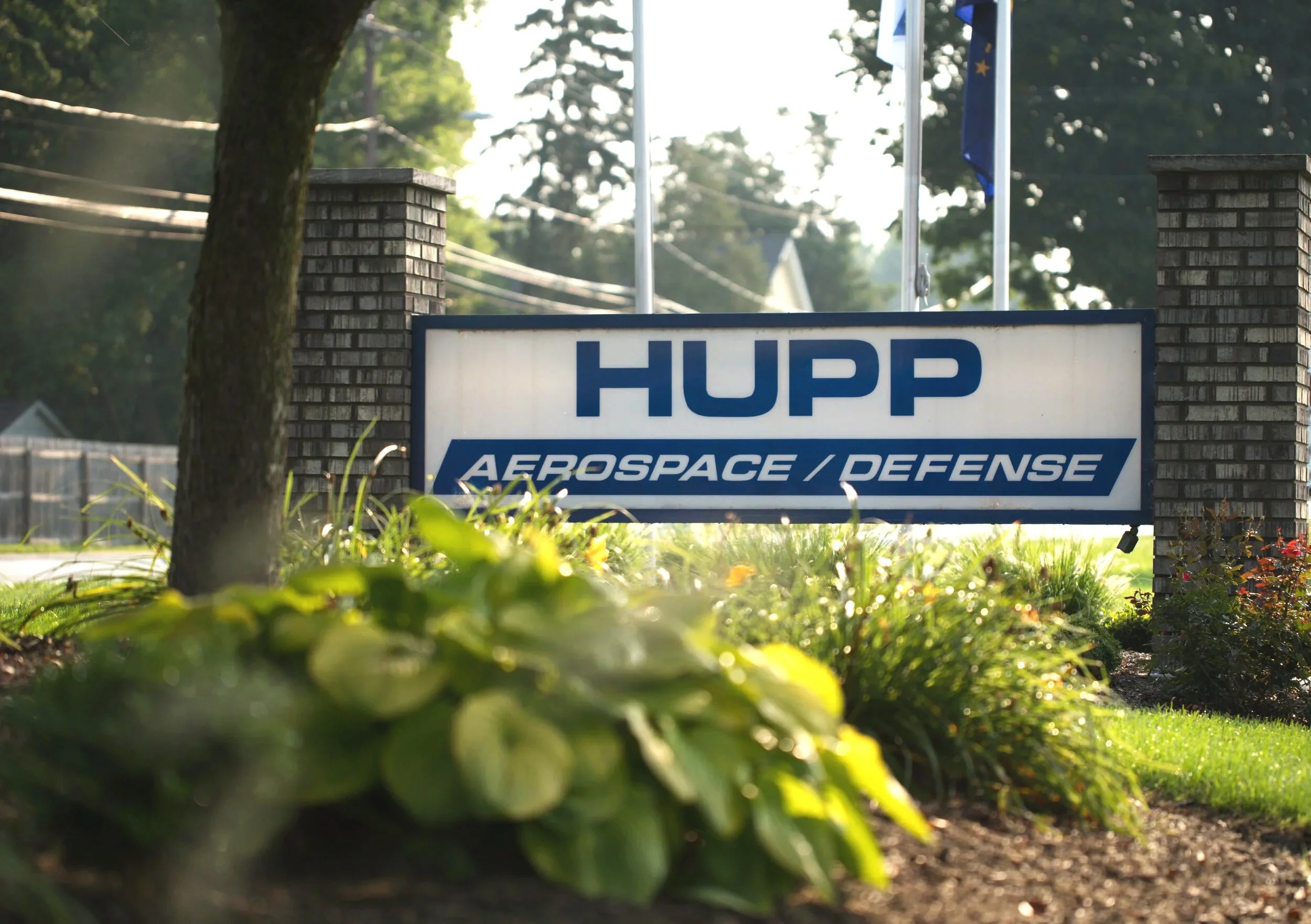 The image shows an outdoor scene with a sign that reads 'HUPP Aerospace/Defense,' supported by brick pillars, surrounded by grass and plants, and trees in the background with flags visible behind the sign.