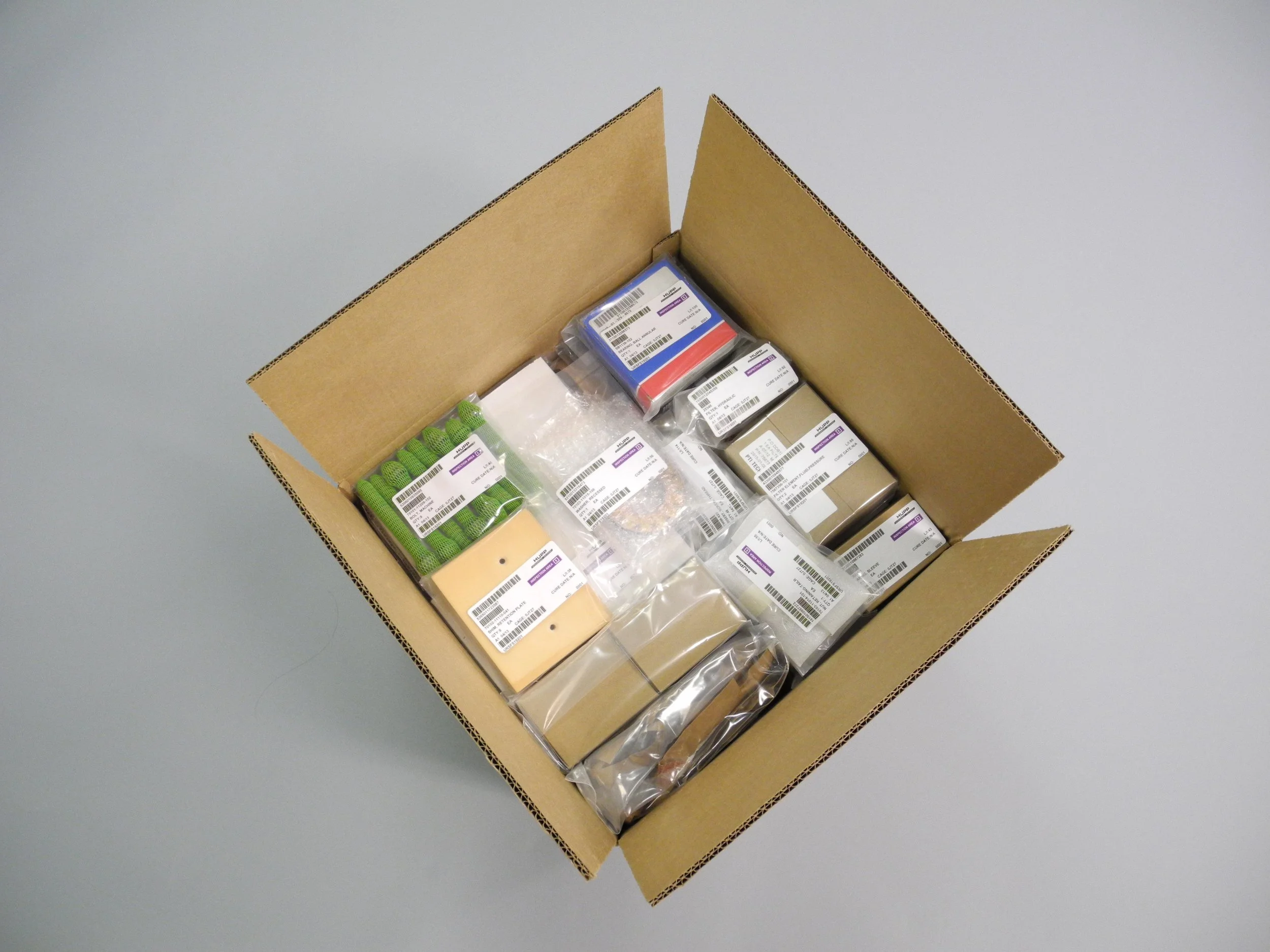 Open cardboard box filled with various sealed component packages, some with barcodes and labels may contain seals, bearings, hardware.