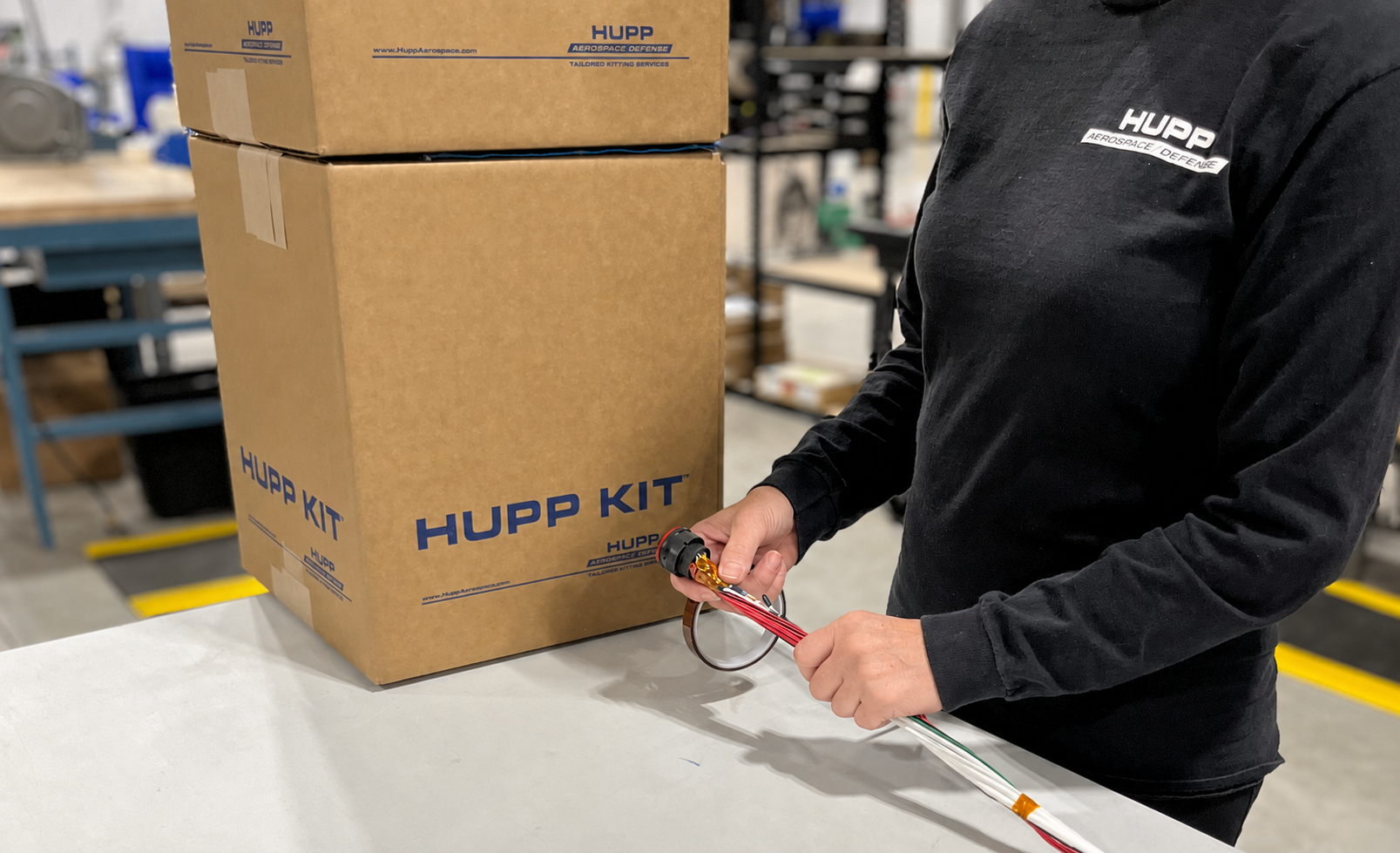 Person working on aerospace wire harness subassembly in a workshop, wearing a black HUPP Aerospace Defense shirt, handling electronic wiring that is pinned into a connector next to a large cardboard box labeled 'HUPP KIT'.