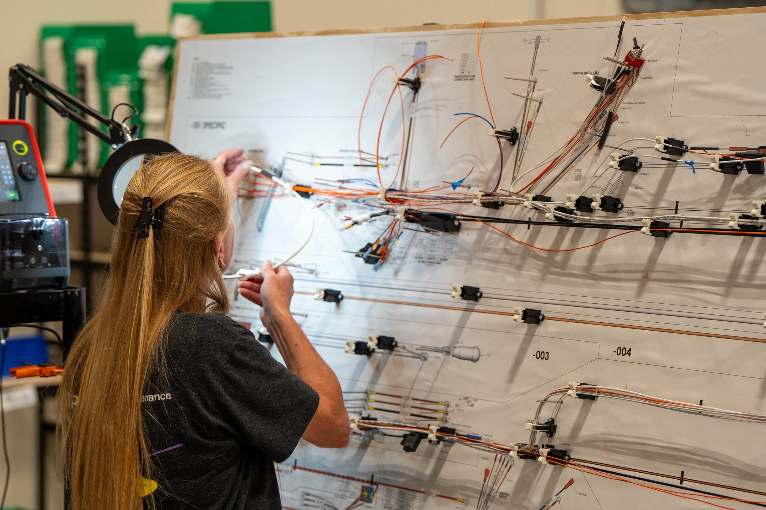 A person working on an electrical wiring harness on a large formboard diagram mounted on a build board, using tools and wearing a black shirt.
