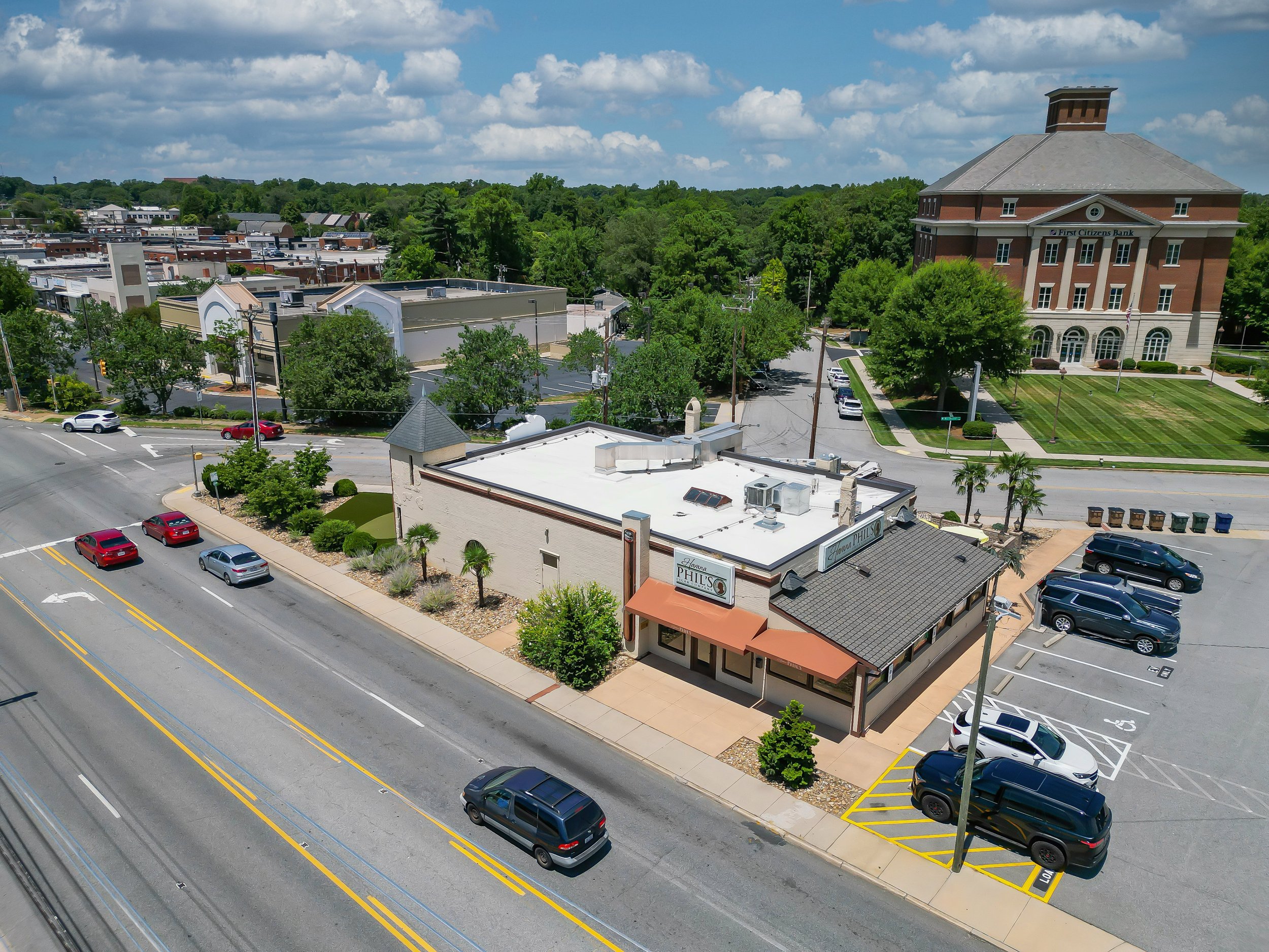 View of a small town with commercial buildings, parking lot, trees, and a large brick building labeled 'First Citizens Bank' under a partly cloudy sky.