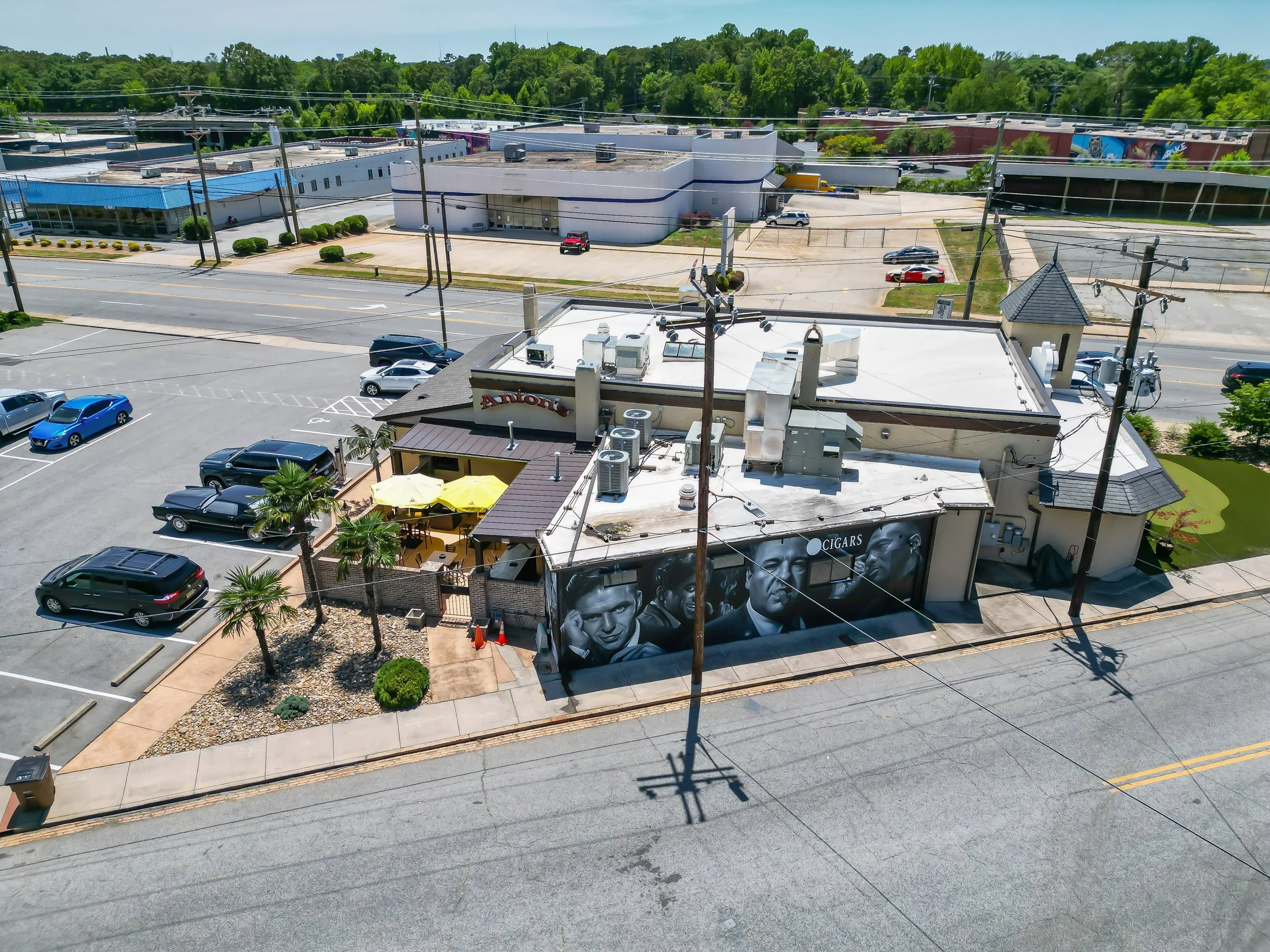 Aerial view of a commercial area with parking lots, a building with artwork of people on the wall, and streets with cars. In the background, there are forested hills.