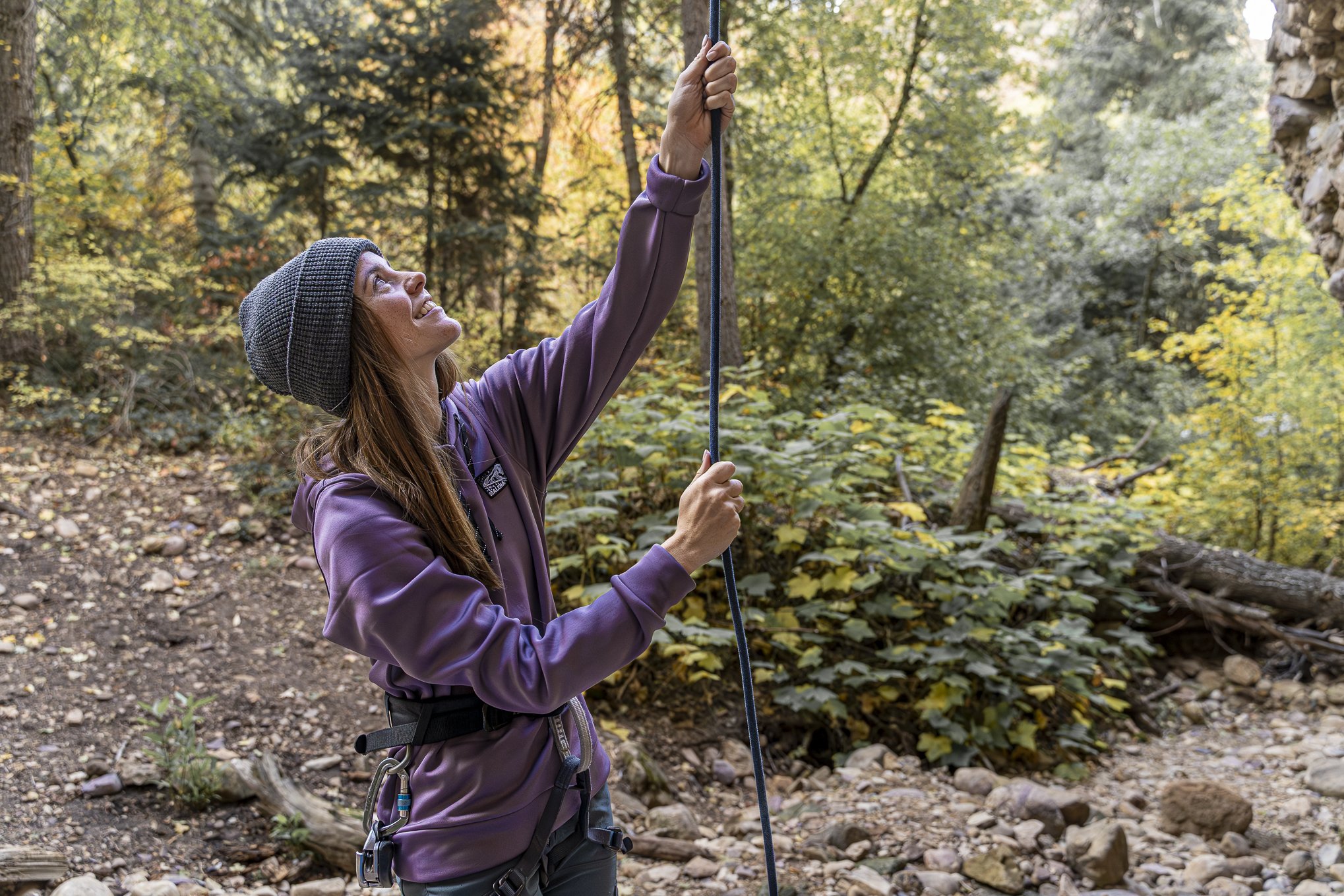 A woman in outdoor clothing and a beanie hat holding a hiking pole in a forest with fall foliage.