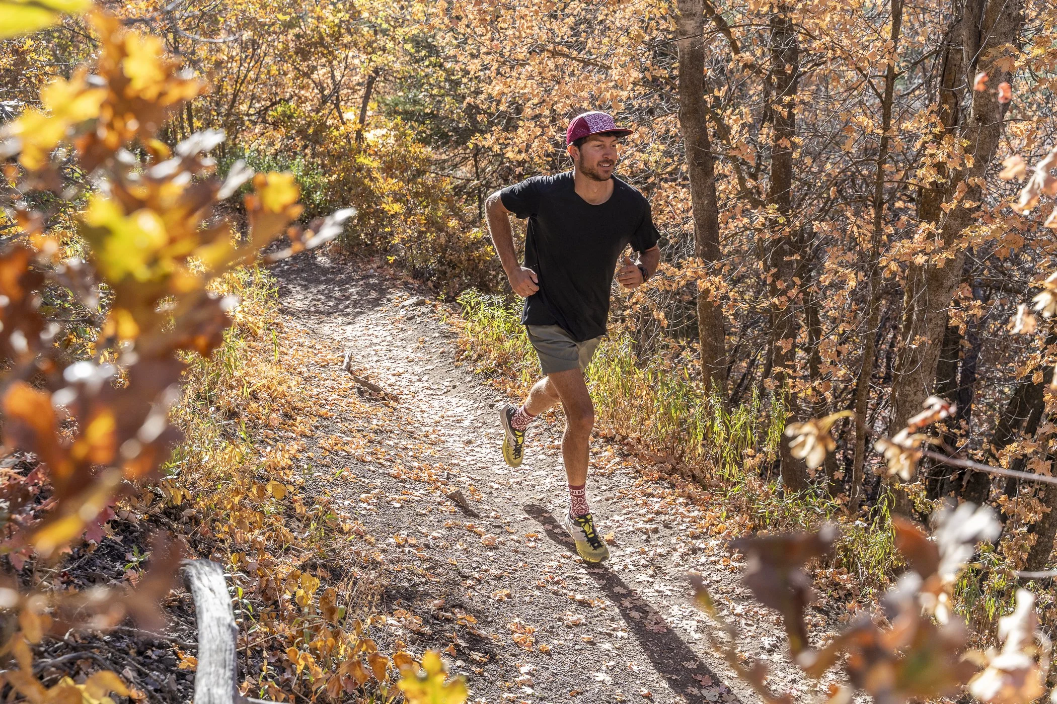 A man running on a dirt trail through a forest with autumn-colored leaves during daytime.