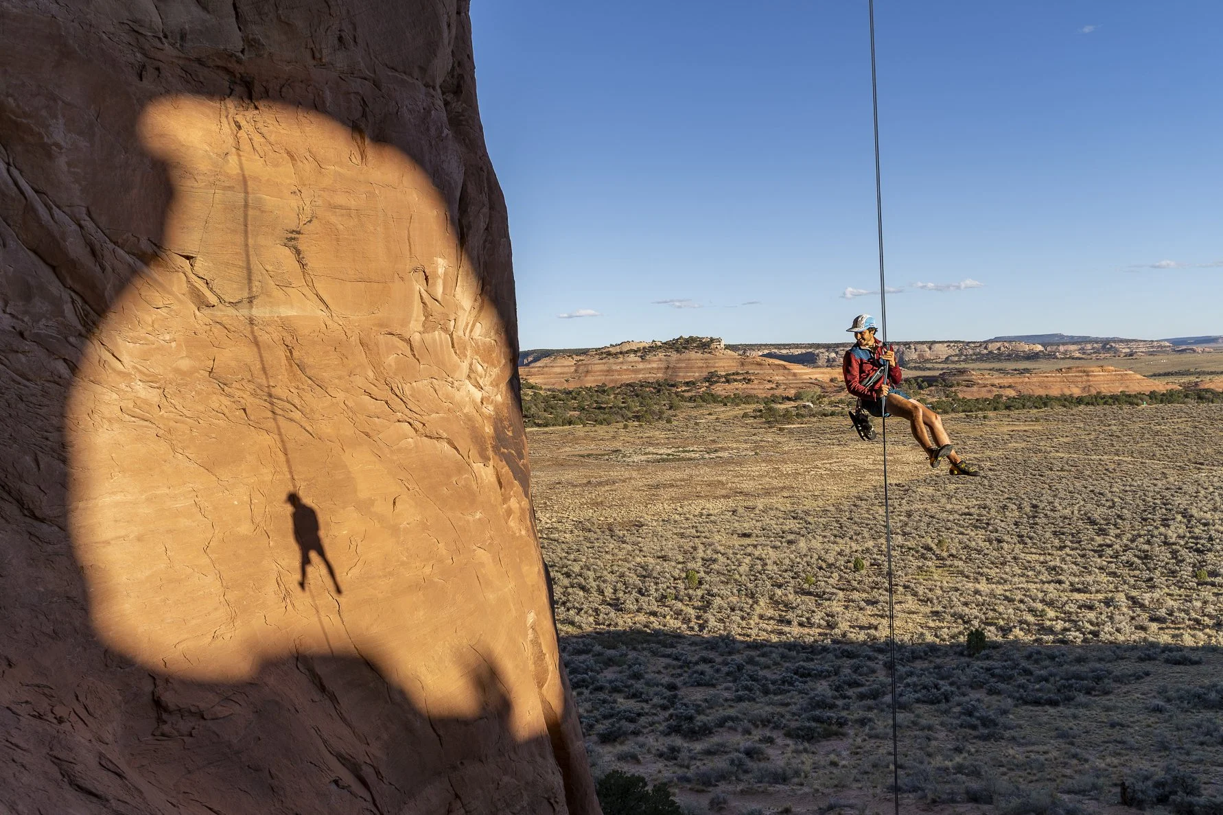A person wearing a helmet and harness is swinging on a rope in the desert landscape, with the shadow of their swing projected onto a red rock face behind them.