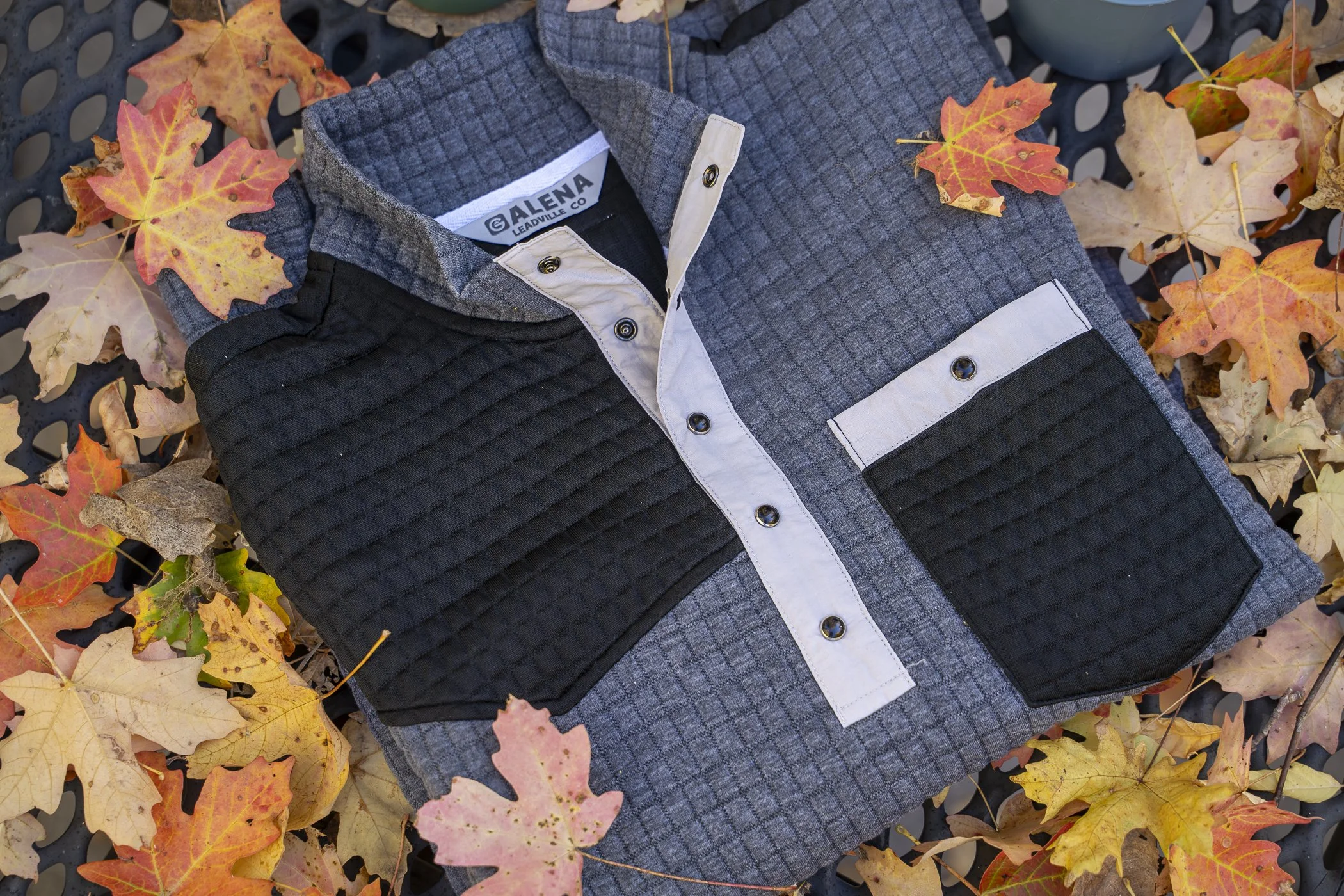 A folded button-up shirt with a gray and black pattern, placed on autumn leaves, with a blue ionized surface.