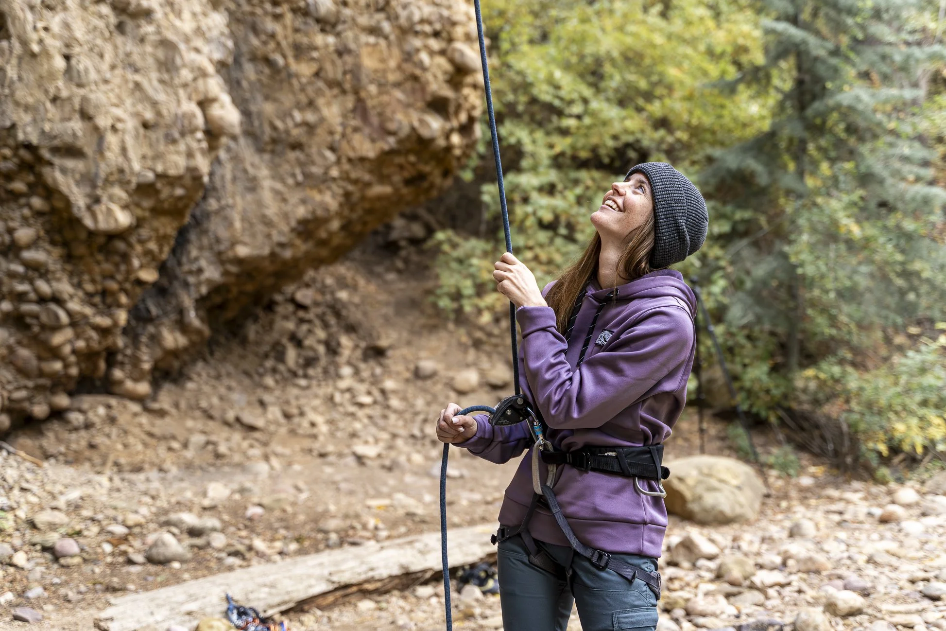 A woman in outdoor clothing and a beanie, holding a rope and looking up, preparing to rock climb or belay in a forested outdoor setting.