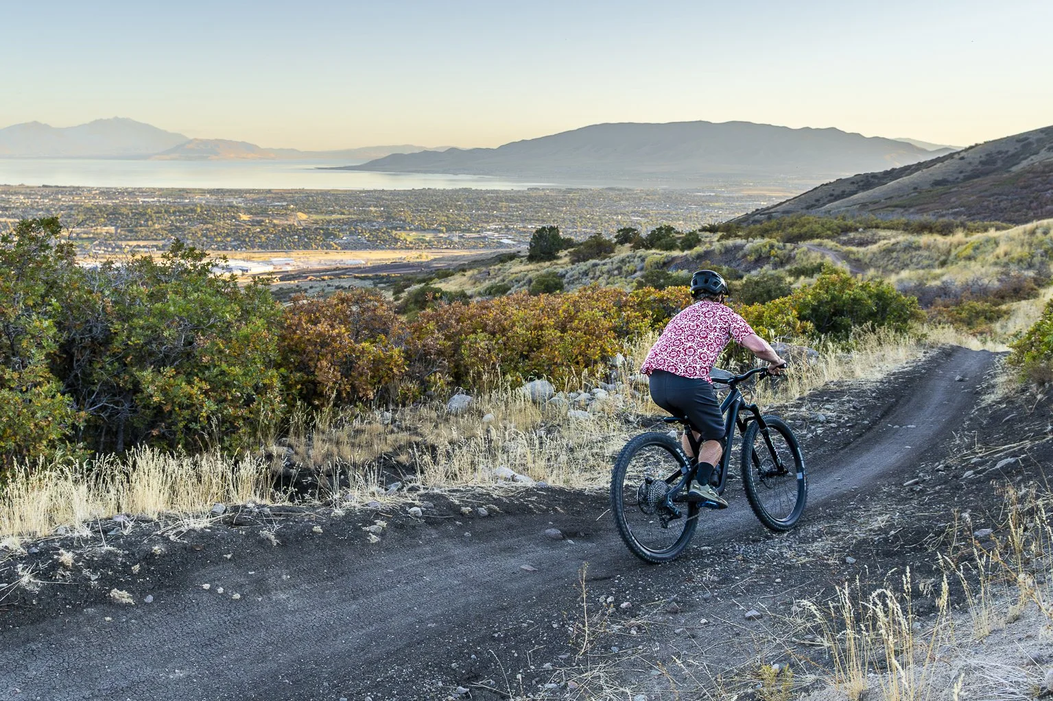 A person riding a mountain bike on a dirt trail with a scenic landscape of mountains, lake, and city in the background, during daytime.