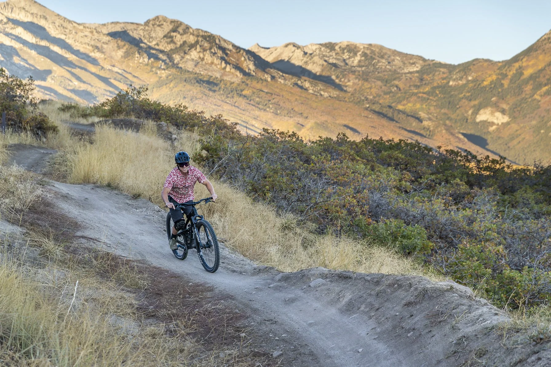 A person riding a mountain bike on a dirt trail in a mountainous landscape with dry grass and green shrubbery, mountains in the background, during daytime.