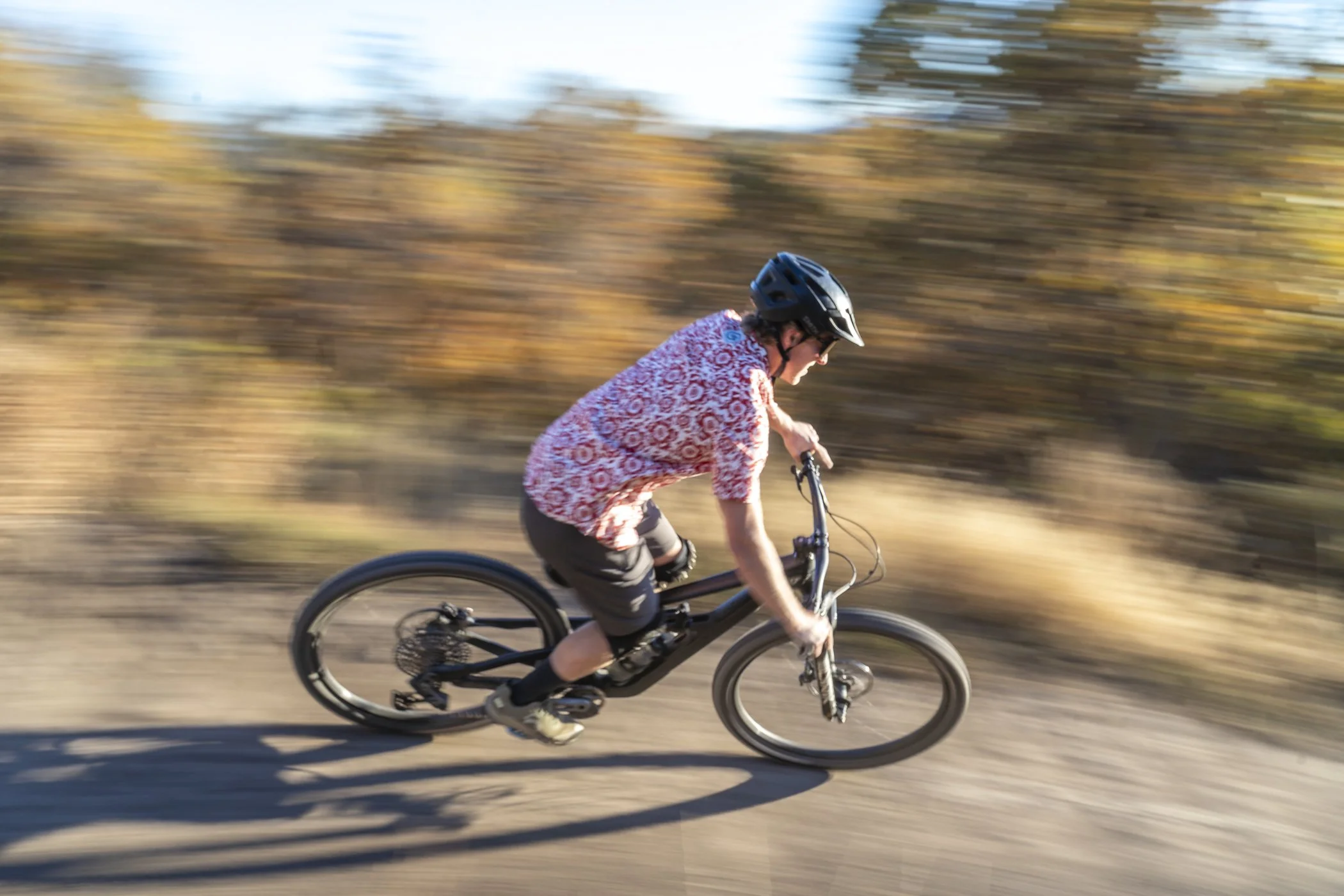 A person wearing a black helmet and a patterned red and white shirt is riding a mountain bike on a dirt trail. The background features a blurred landscape of trees and foliage, indicating motion and speed.