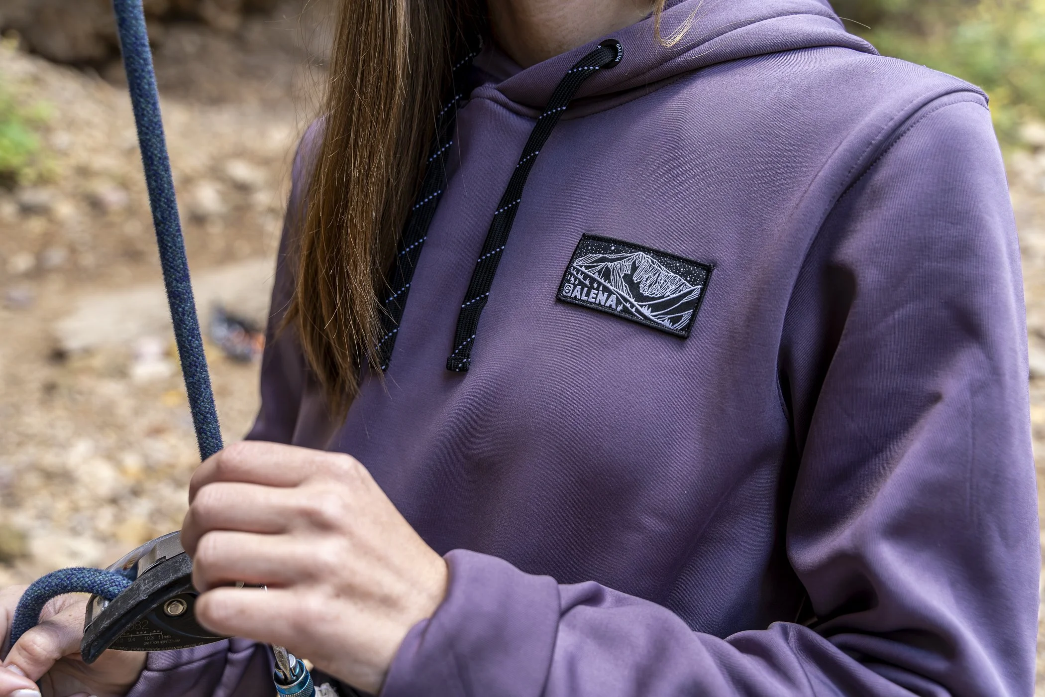 Close-up of a person wearing a purple hooded jacket with a mountain patch labeled 'Galena', holding climbing gear and a rope, outdoors in a natural setting.