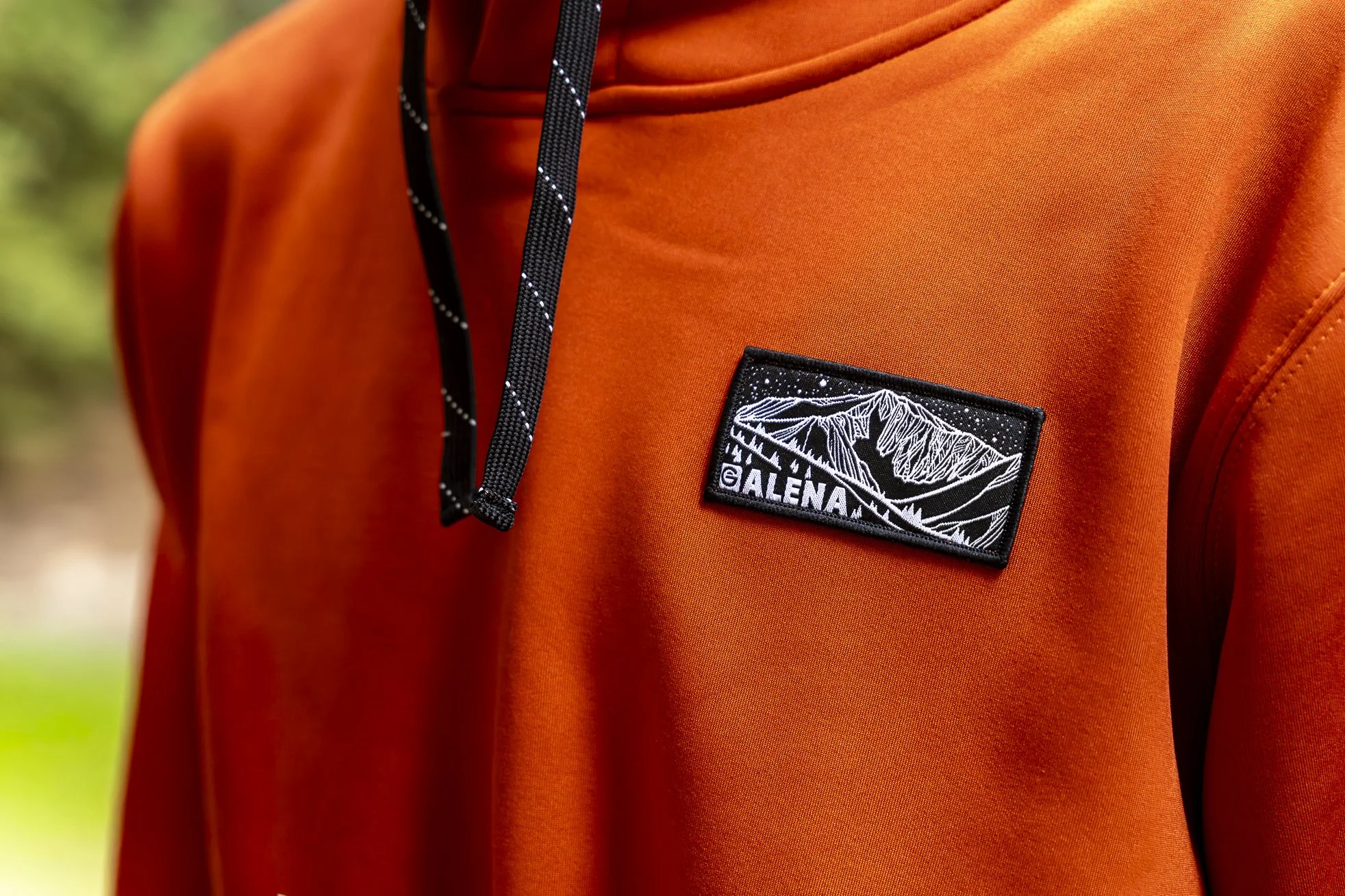 Close-up of an orange outdoor jacket with a black and white patch displaying mountain peaks and the brand name 'GALENA'.