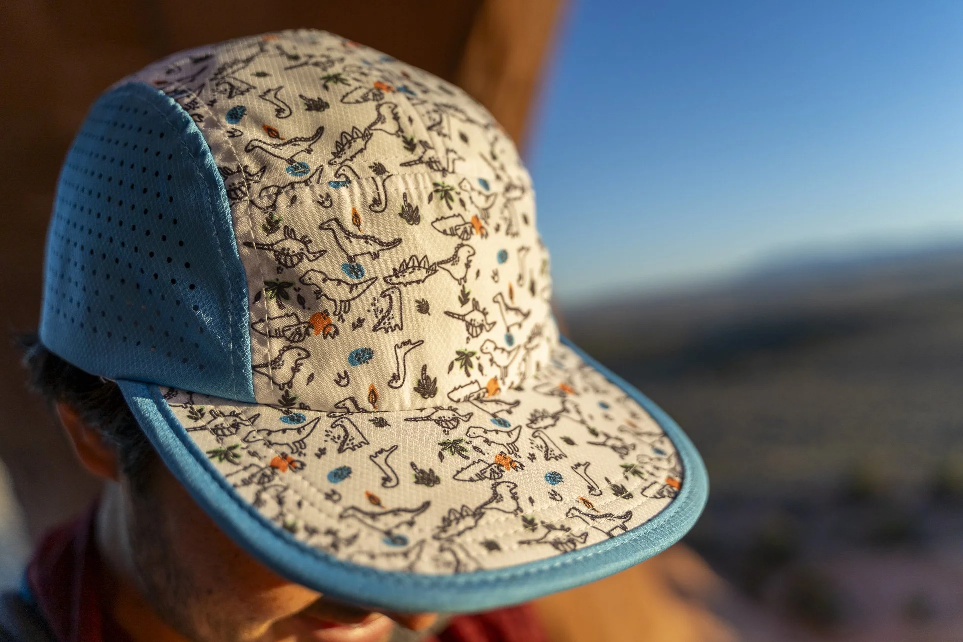 A person wearing a white and blue dinosaur-patterned cap outdoors during sunset or sunrise, with ocean and sky in the background.