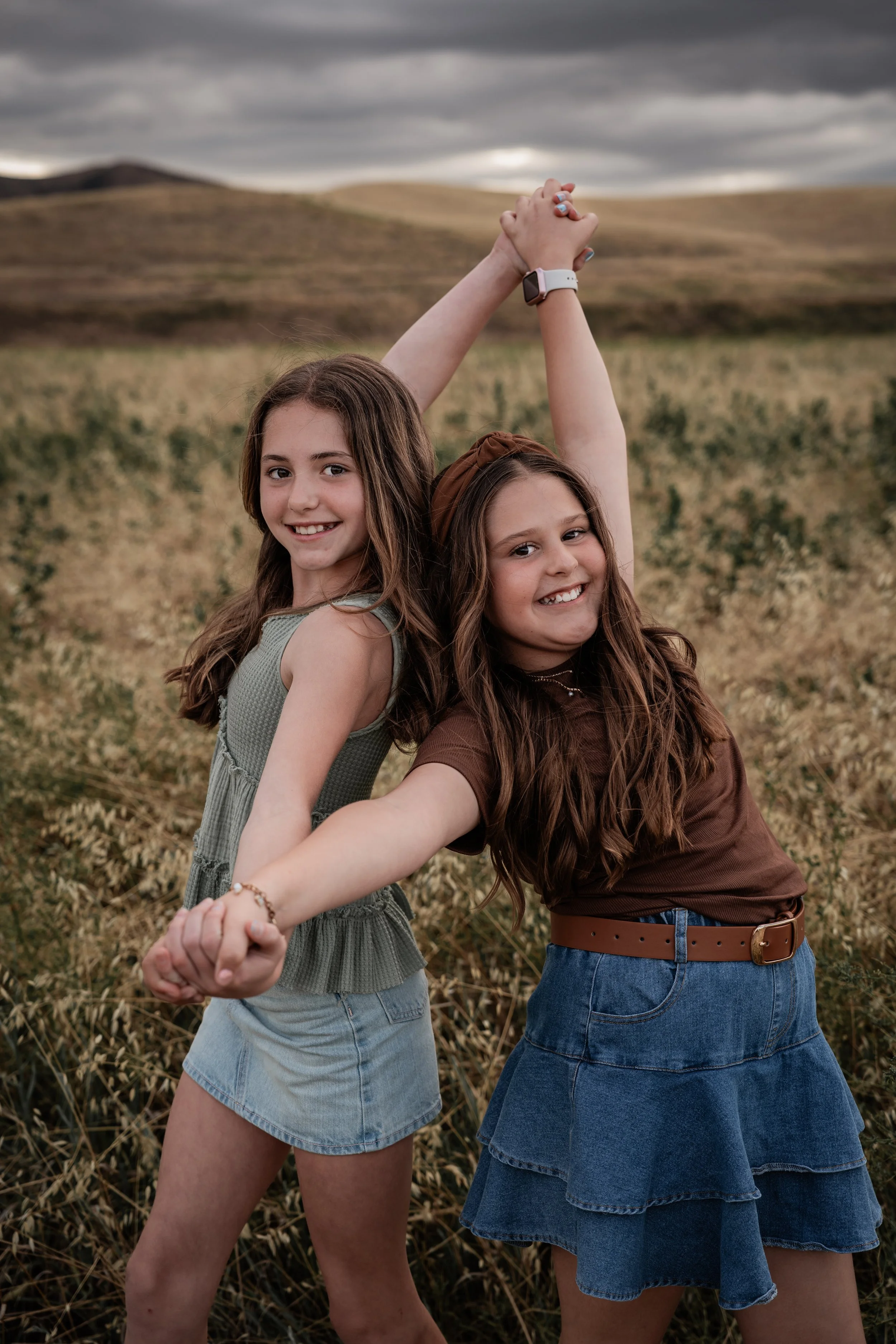 Two girls holding hands and posing while standing in an open field. Photo taken by family photographer, Mikaela Tosta Photography in Patterson, CA.