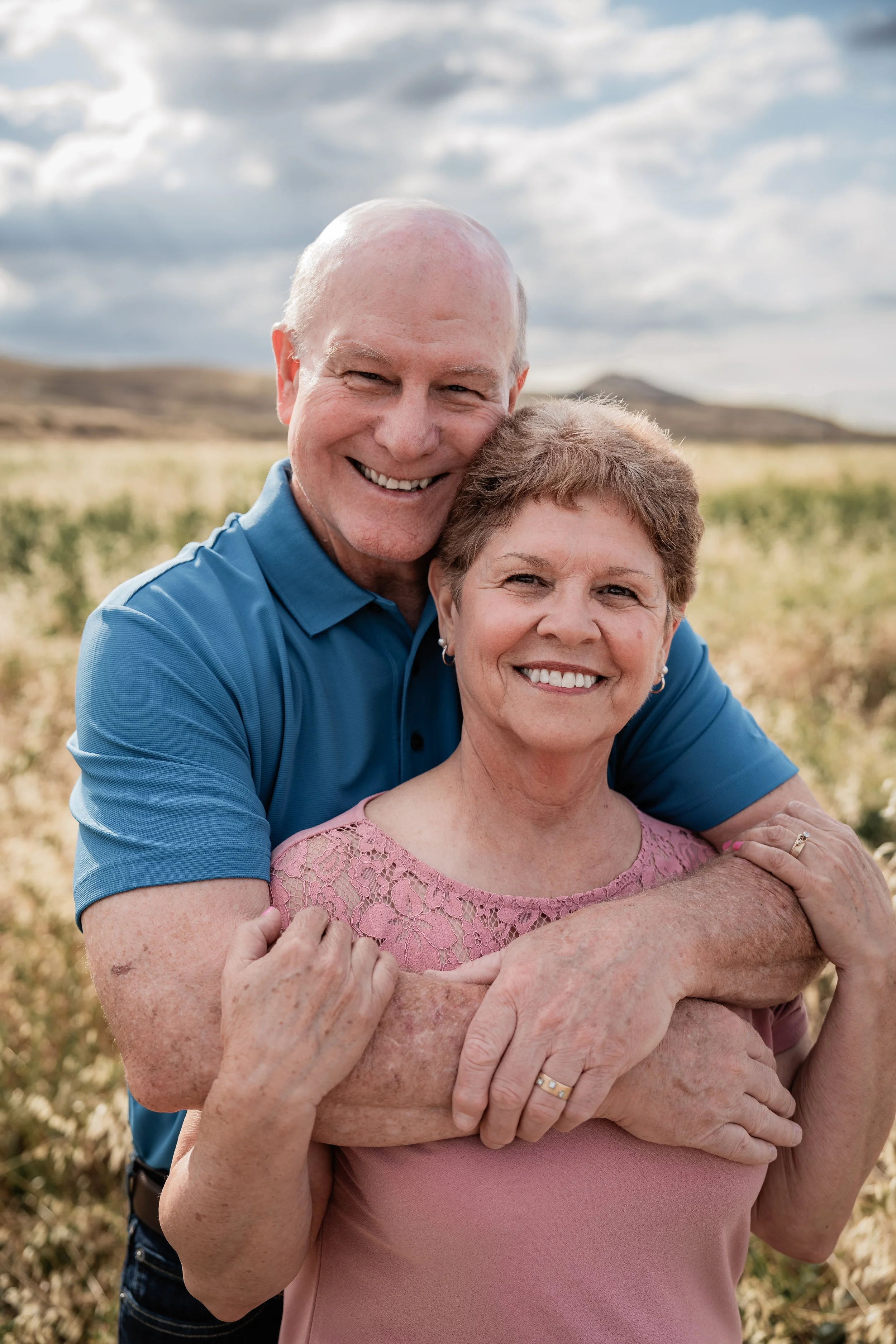Couple hugging while looking at the camera standing in an open field. Photo taken by family photographer, Mikaela Tosta Photography in Patterson, CA.