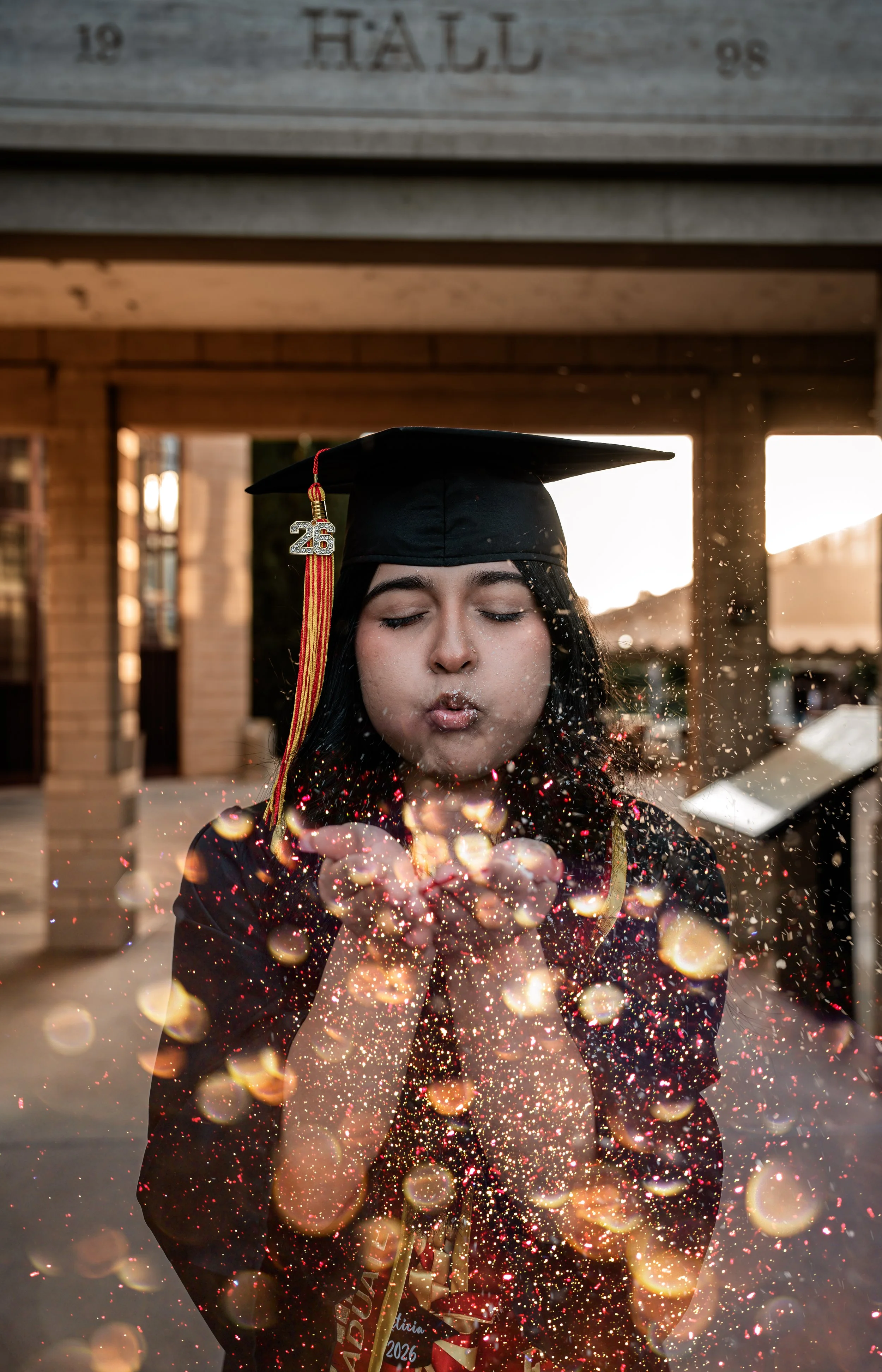 Photo of a Stanislaus State Graduate in front of DBH blowing glitter out of her hand. Photo taken in Turlock, CA by senior photographer and grad photographer Mikaela Tosta Photography.