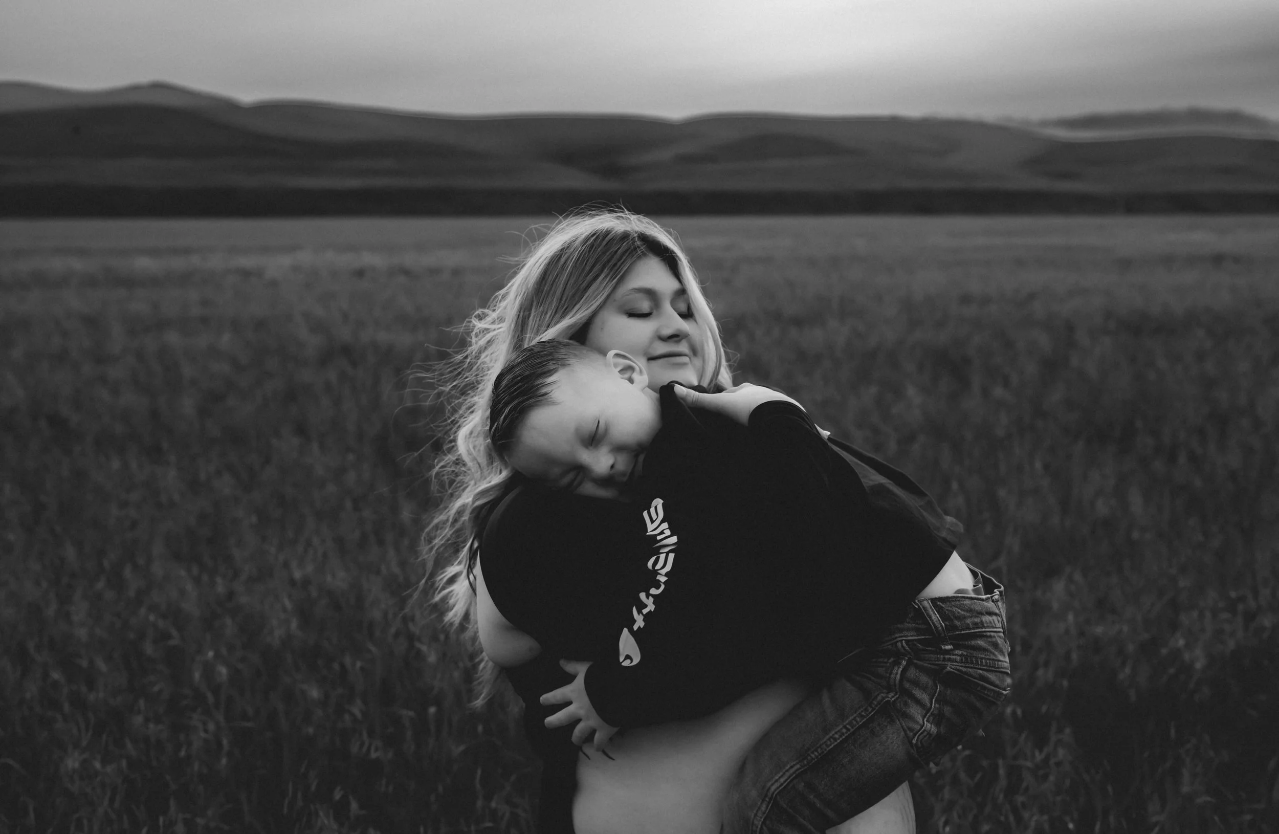 Black and white photo of pregnant mother snuggling with her son in an open field. Photo taken in Patterson, CA by Mikaela Tosta Photography.