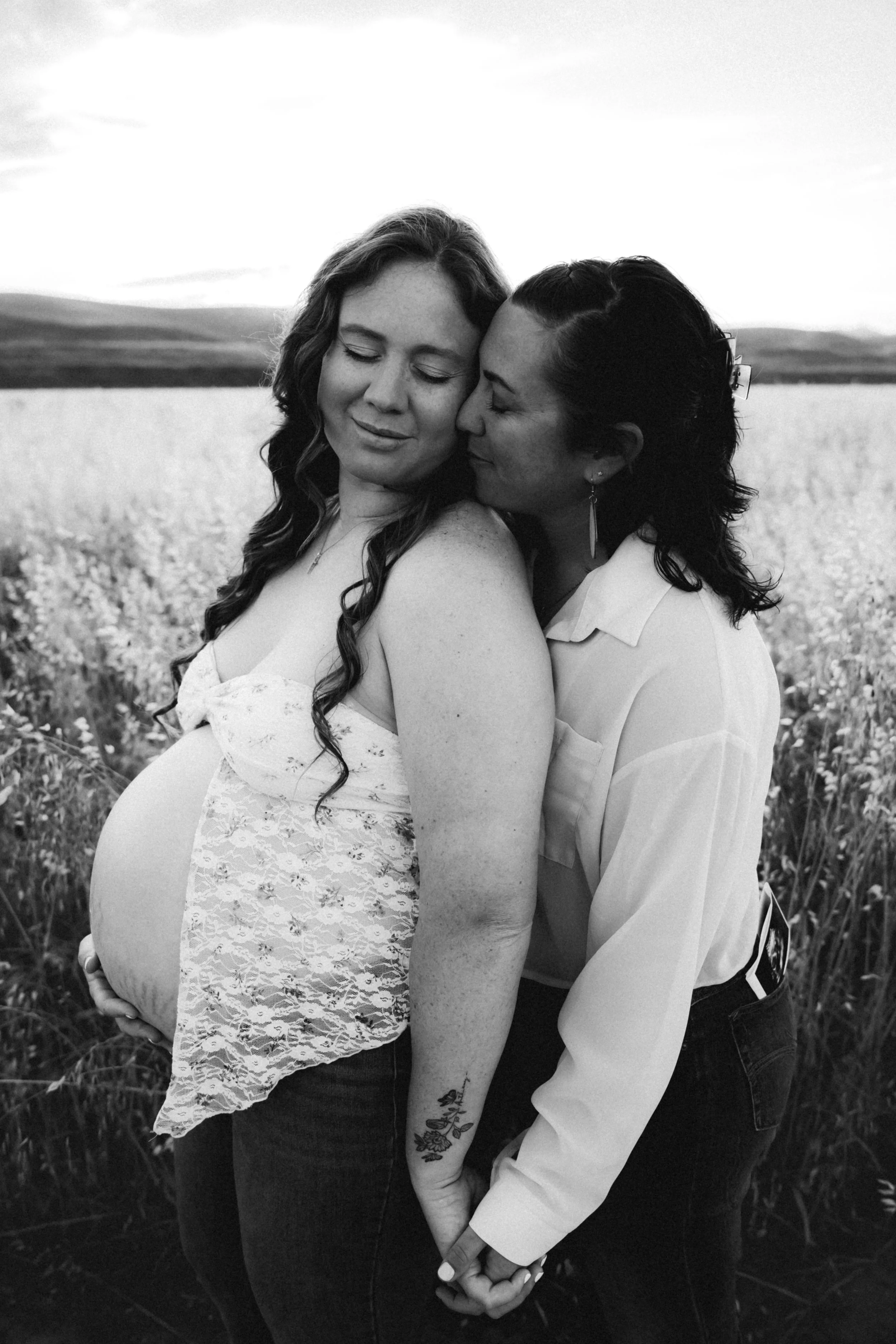 Black and white photo of pregnant mother snuggling with her wife in an open field. Photo taken in Patterson, CA by Mikaela Tosta Photography.