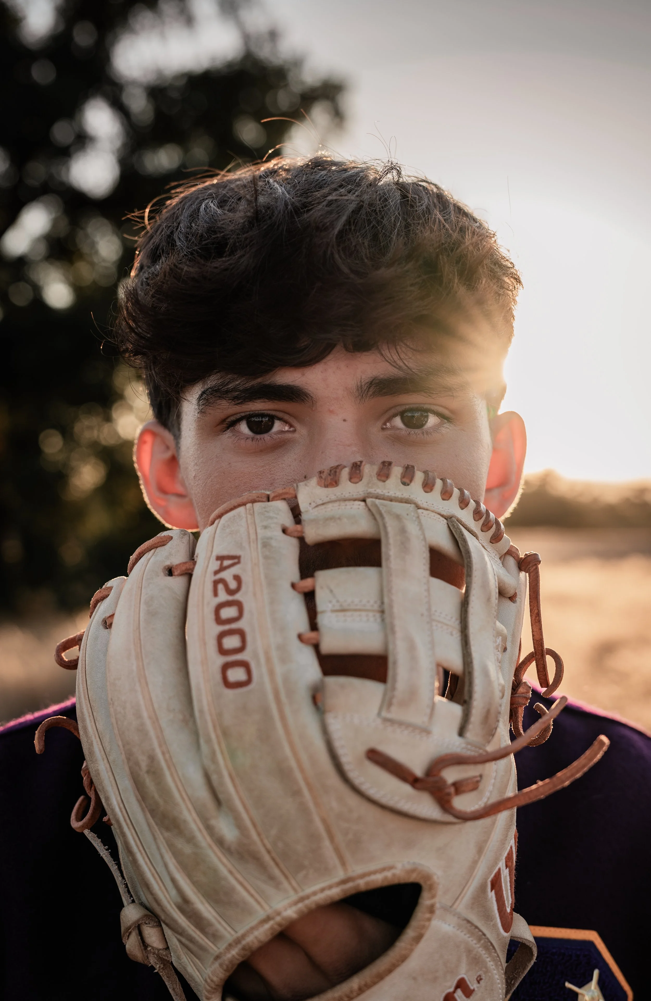 High school senior covering his face with a baseball glove in a field. Photo taken in Newman, CA by Mikaela Tosta Photography