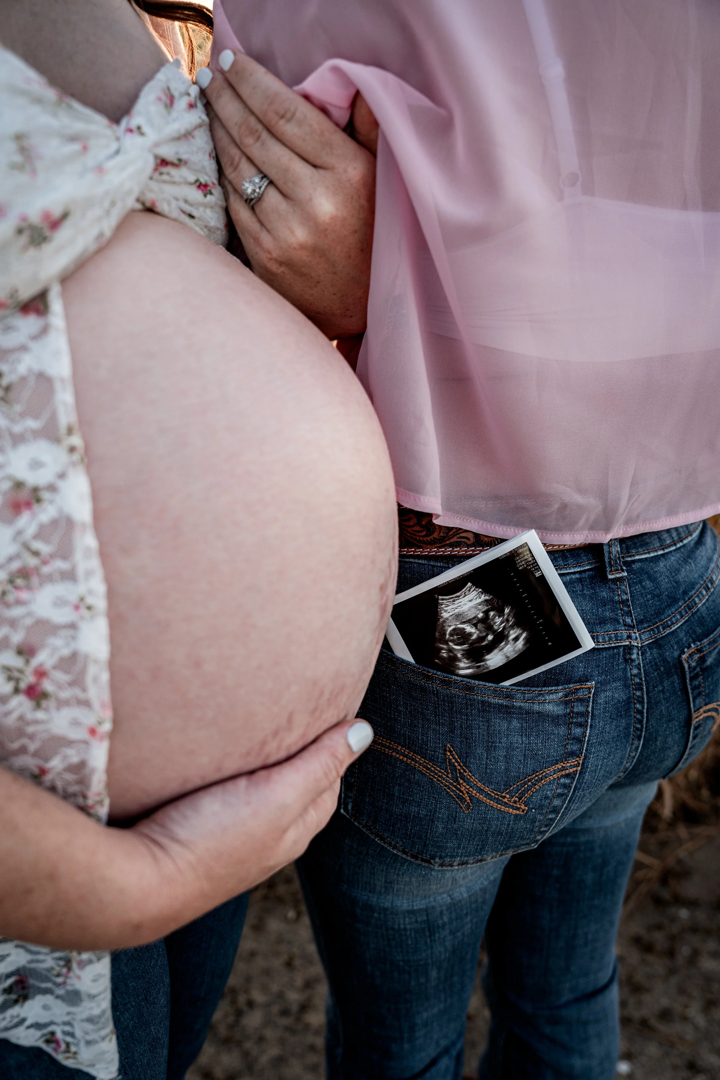 Photo of pregnant mom snuggling her wife with an ultrasound picture being in the wife's back pocket in an open field. Photo taken in Patterson, CA by Mikaela Tosta Photography.