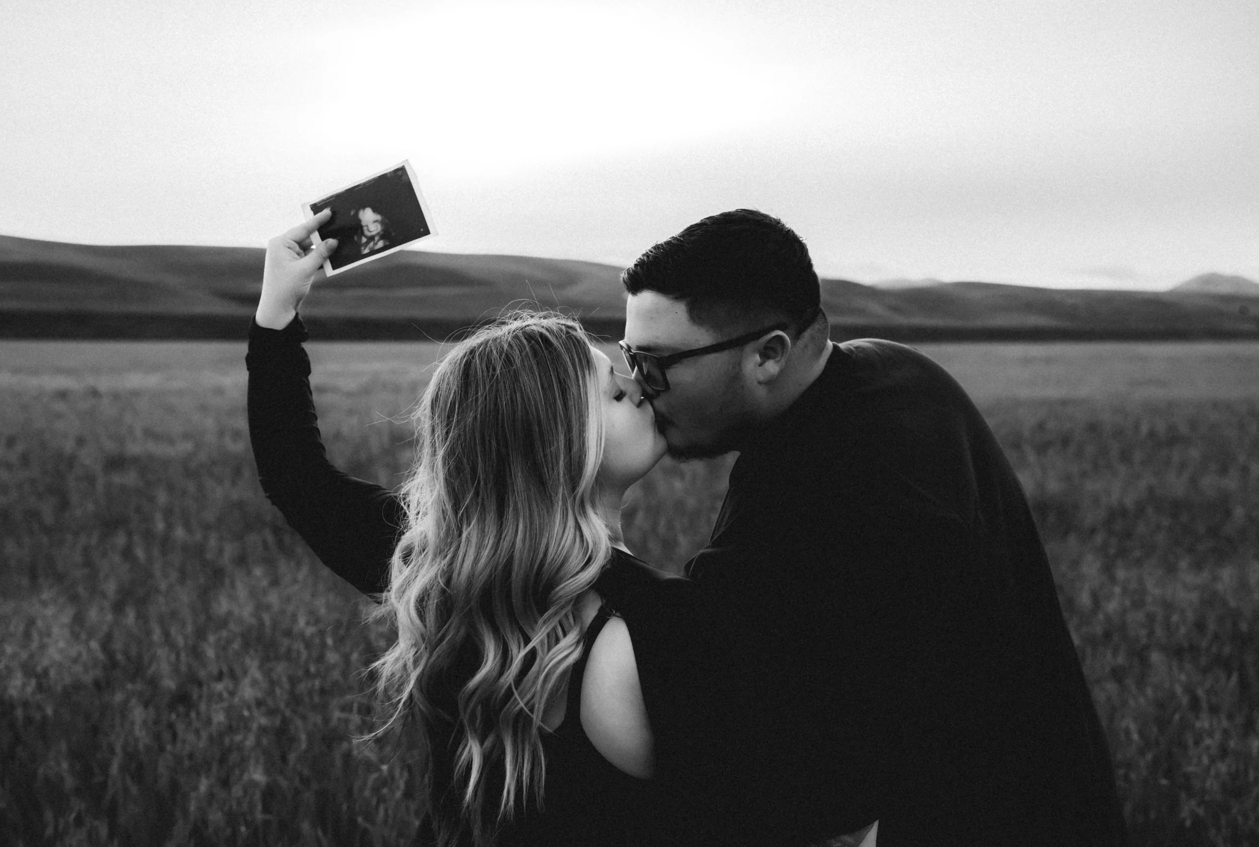Black and white photo of pregnant mother kissing with her partner holding a photo in an open field. Photo taken in Patterson, CA by Mikaela Tosta Photography.