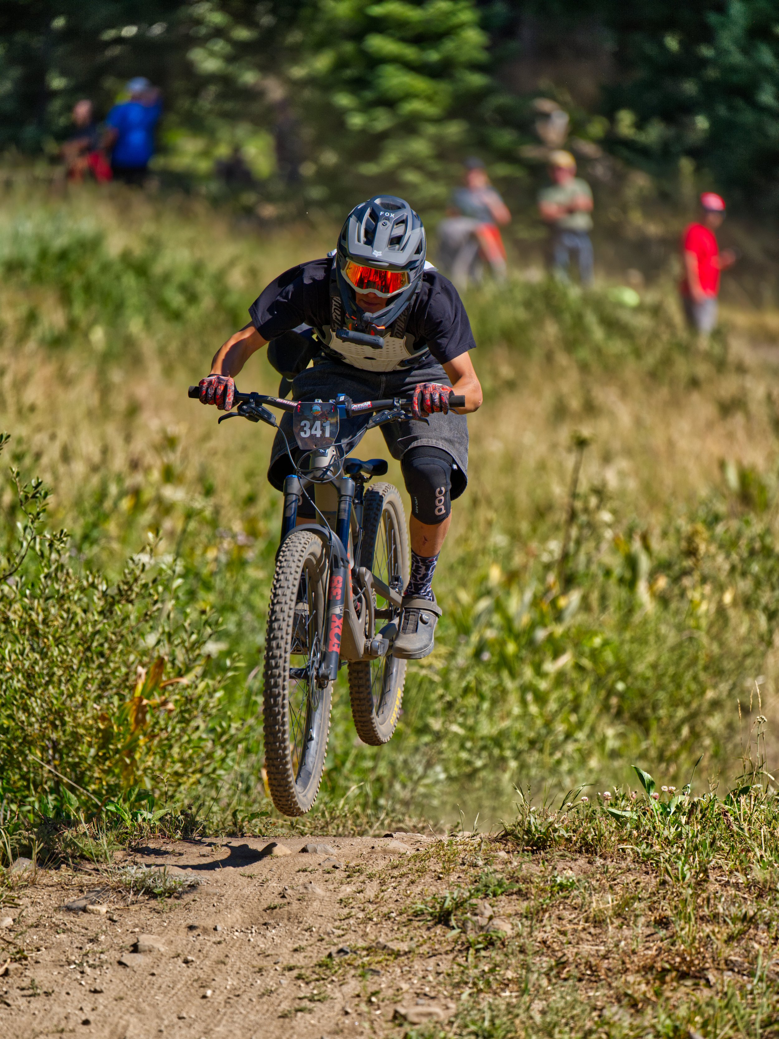 A person in mountain biking gear riding a mountain bike on a dirt trail in a forested area with a group of people in the background.