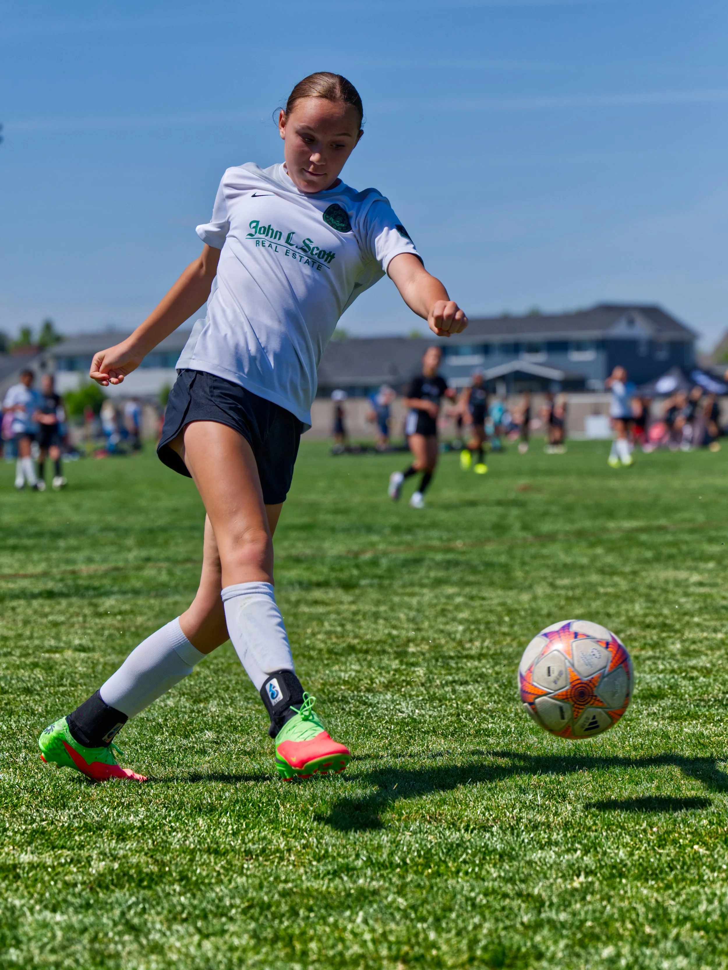 A young female soccer player kicking a soccer ball on a grassy field with other players and houses in the background.